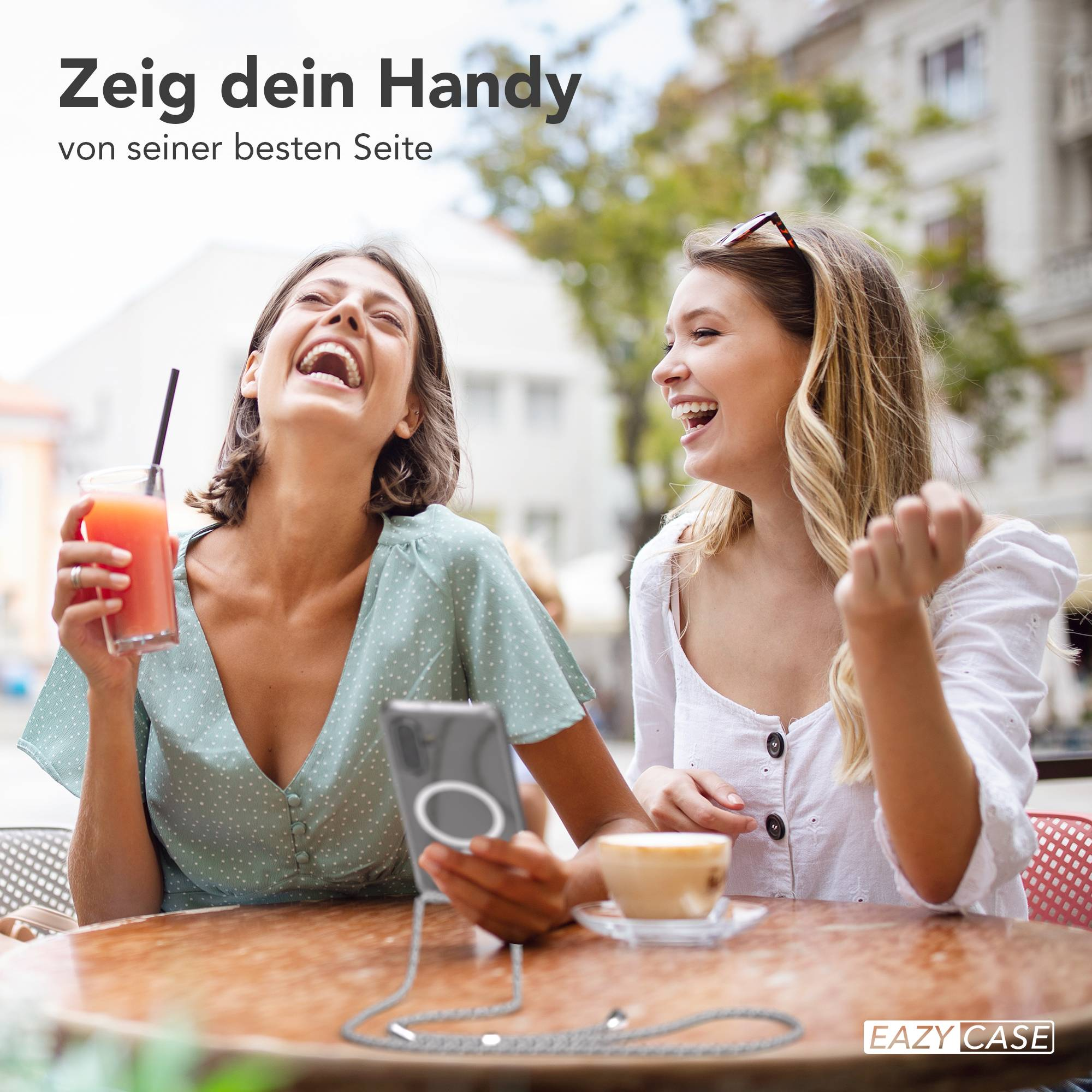 Zwei Frauen lachen und trinken draußen an einem Tisch. Eine hält ein Getränk.