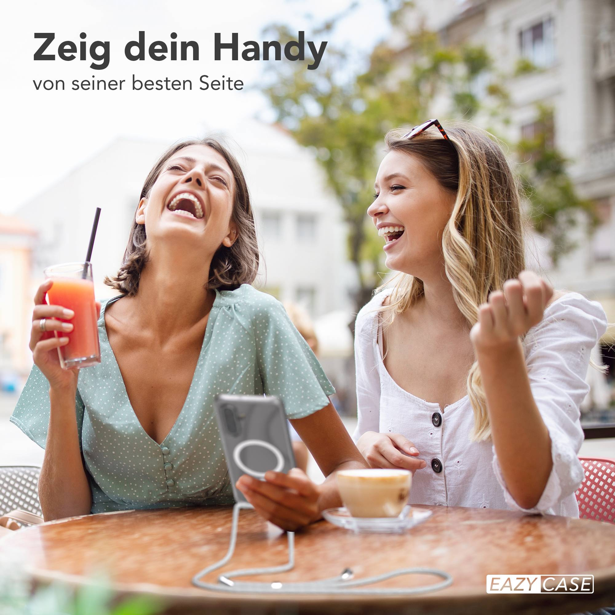 Zwei lachende Frauen sitzen draußen, eine hält ein Getränk, die andere ein Handy.