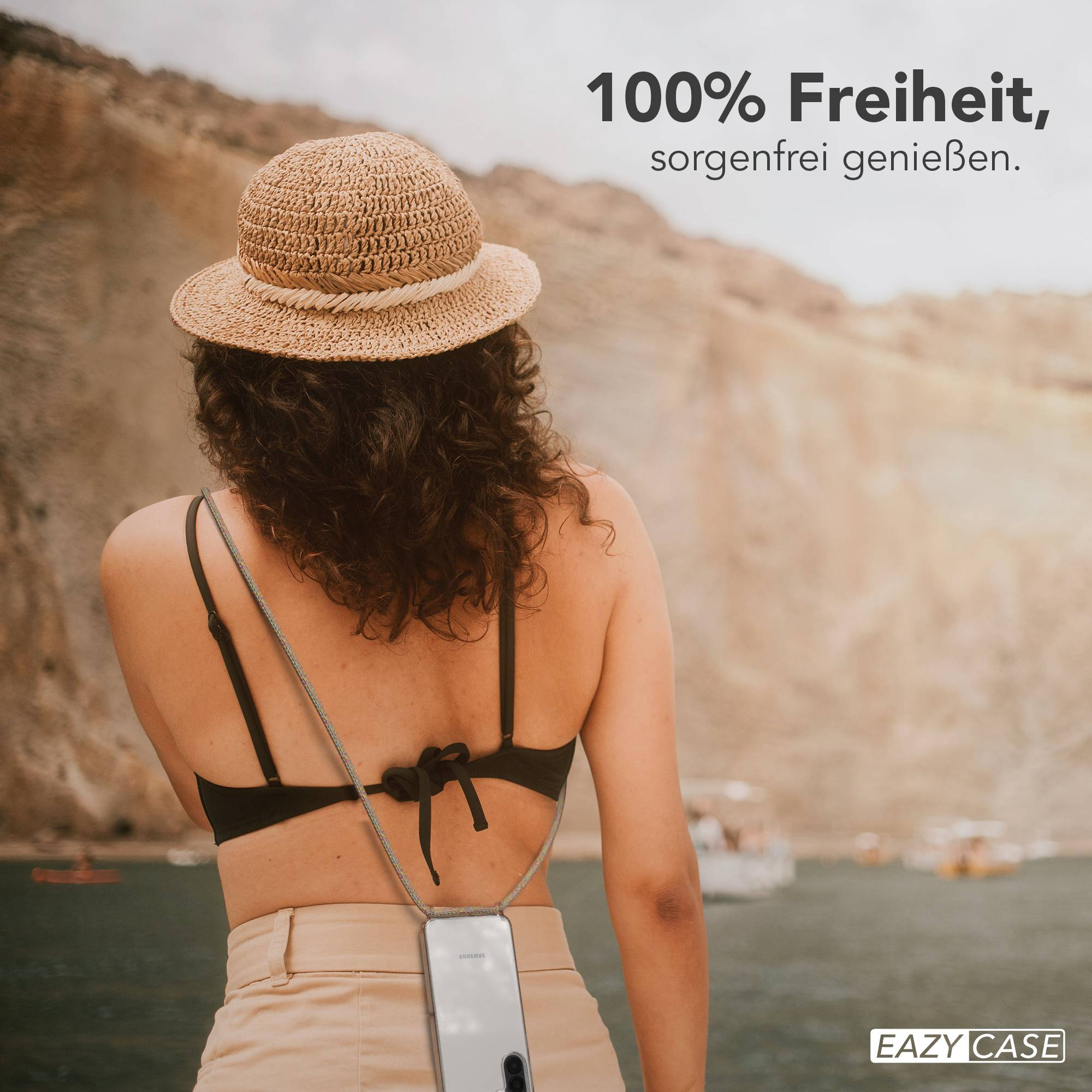 Frau mit Strohhut und Handy-Halterung am Strand bei den Bergen.