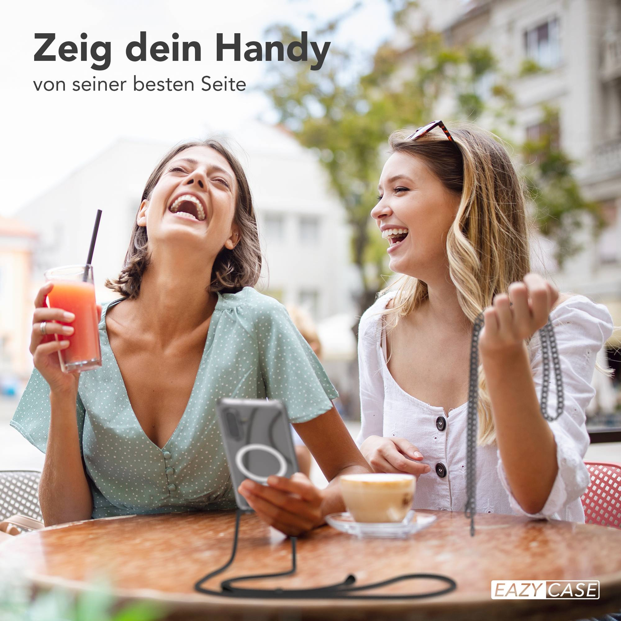 Zwei Frauen lachen draußen an einem Tisch mit Getränken und einem Handy.