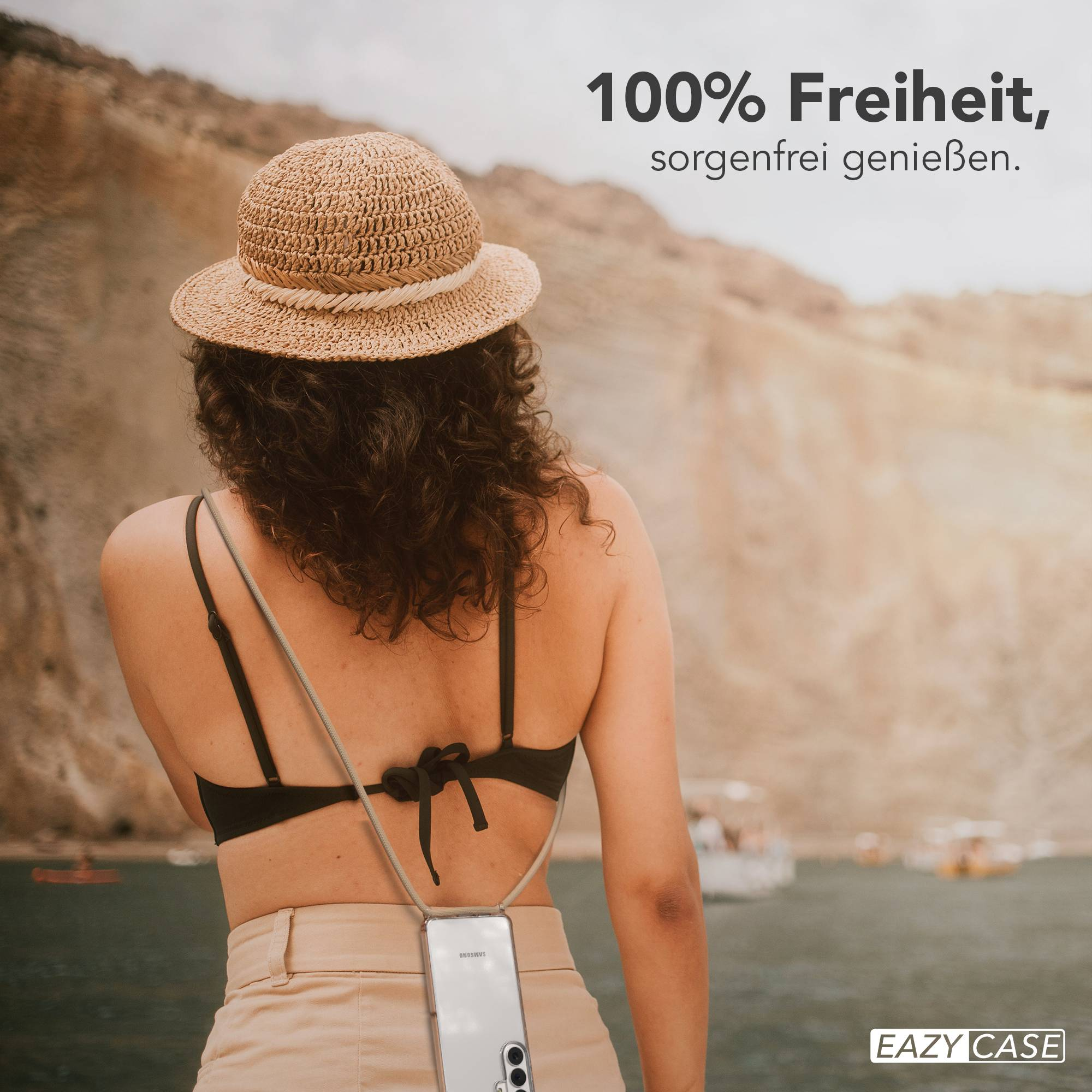 Frau mit Strohhut am Meer, Handy mit Trageband, Boote im Hintergrund.
