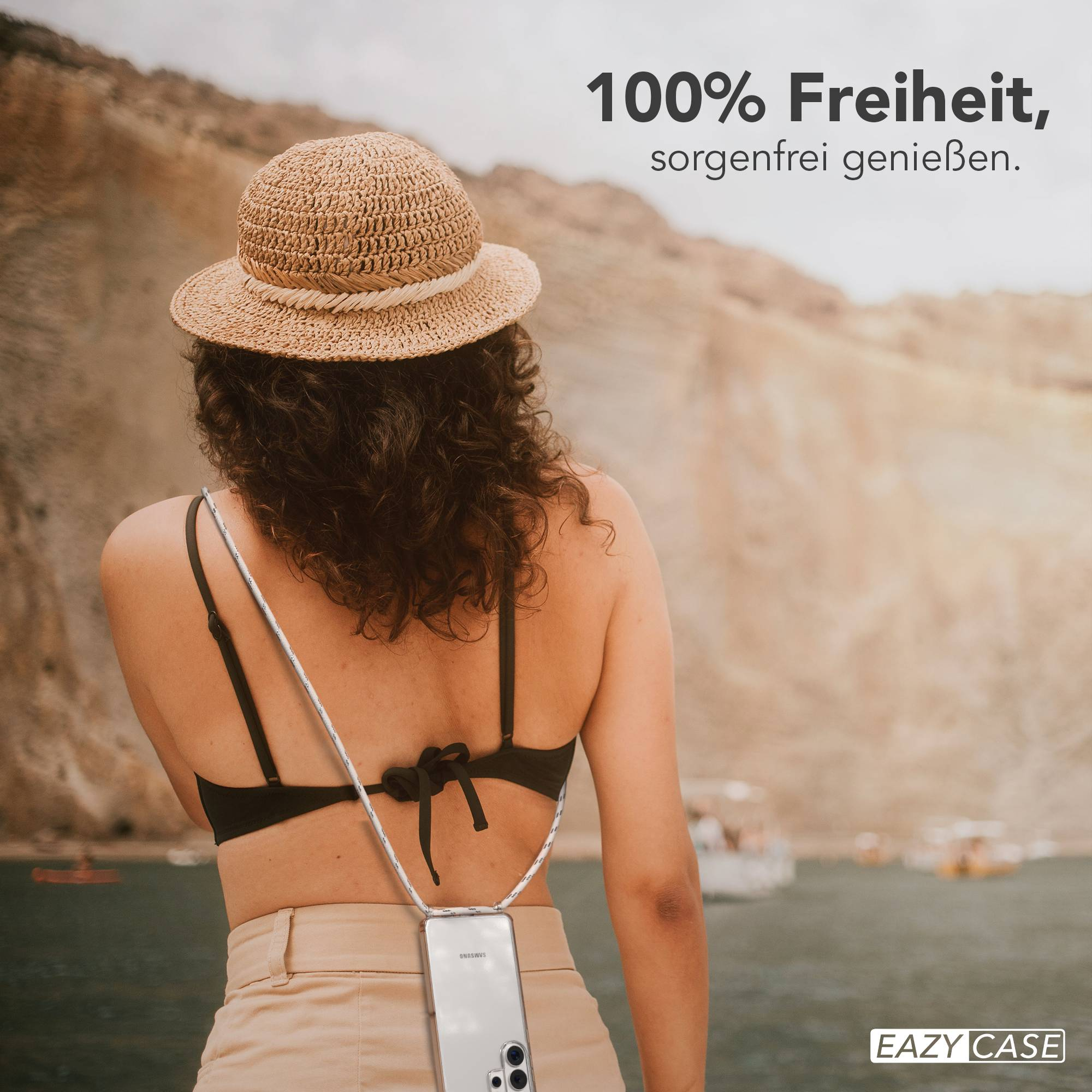 Frau mit Strohhut und Bikini-Oberteil, Handy am Halsriemen, am Wasser.