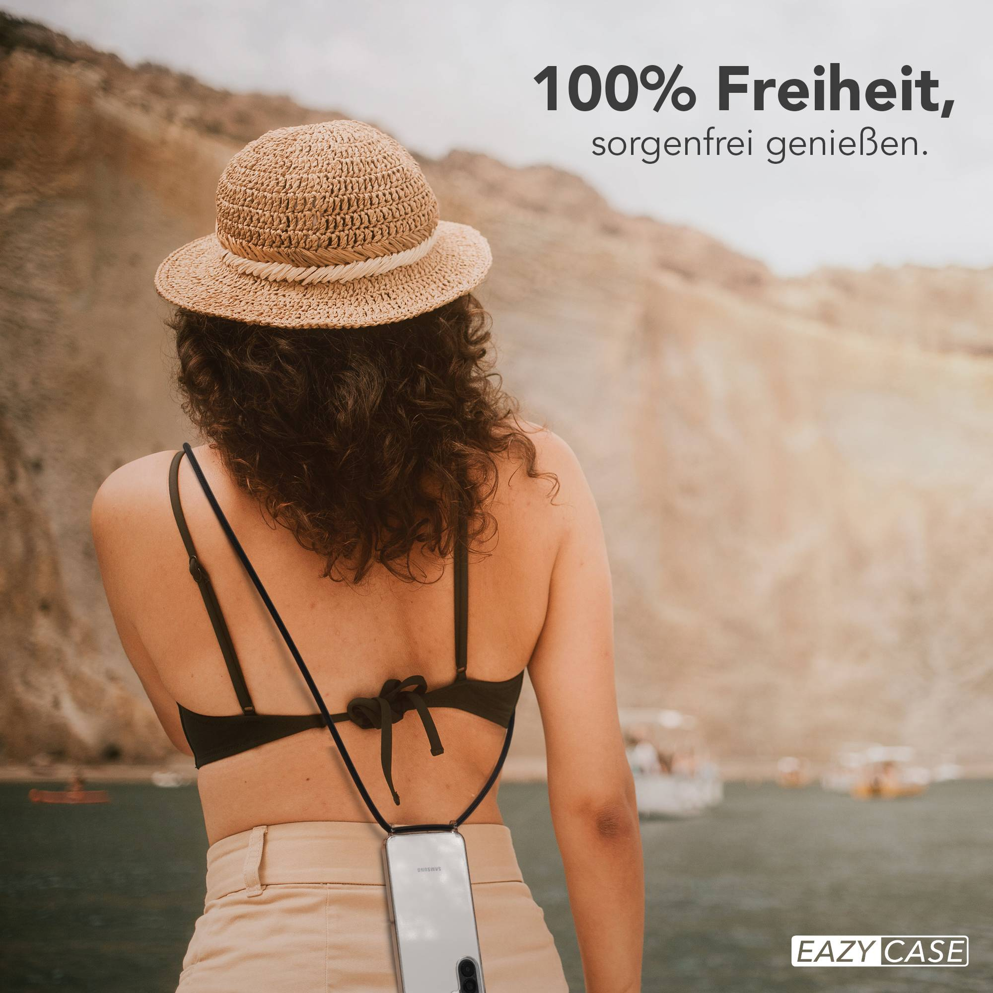Frau mit Strohhut und Handy-Halterung am Strand mit Booten im Hintergrund.
