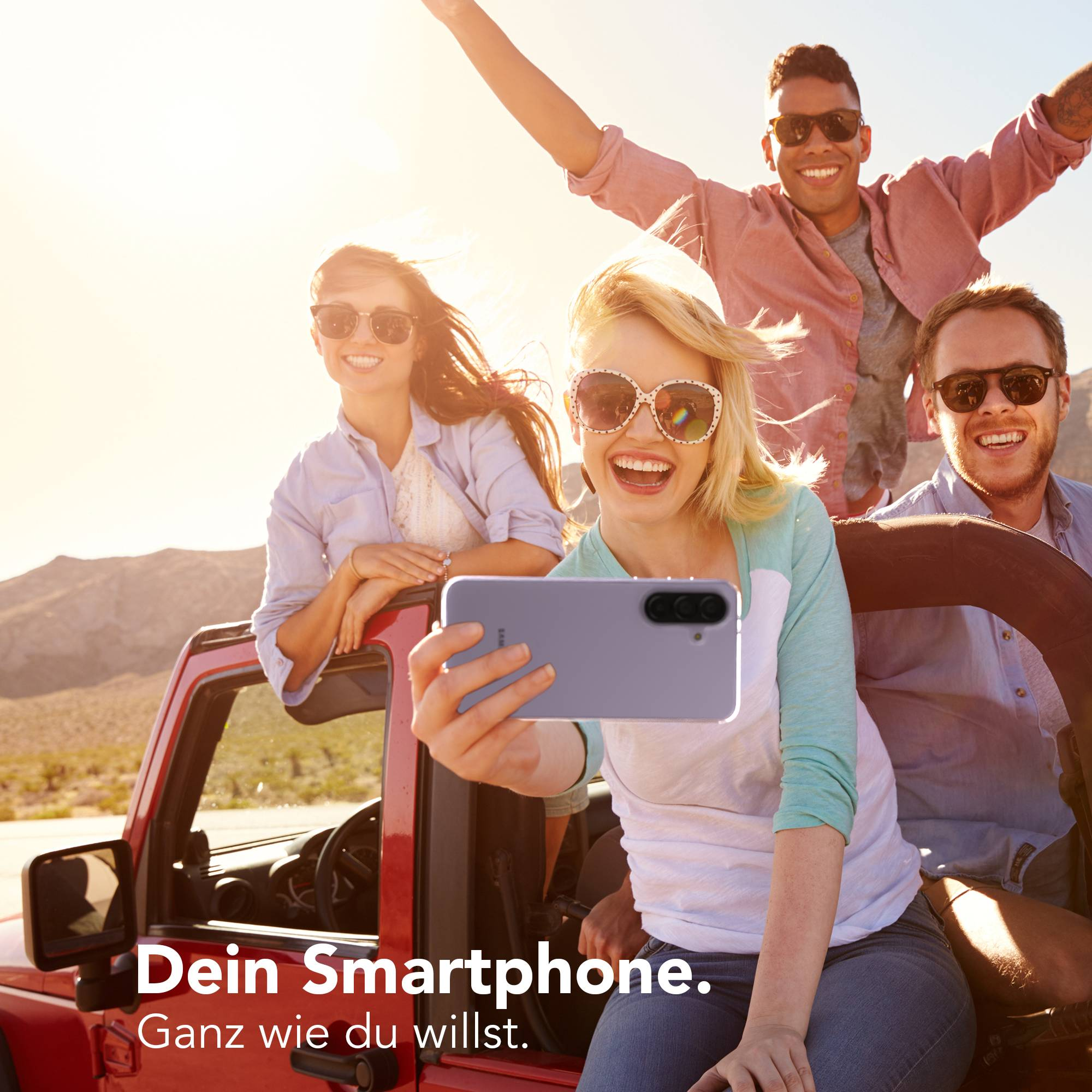 Vier Freunde machen ein Selfie in einem roten Jeep in sonniger Wüste.