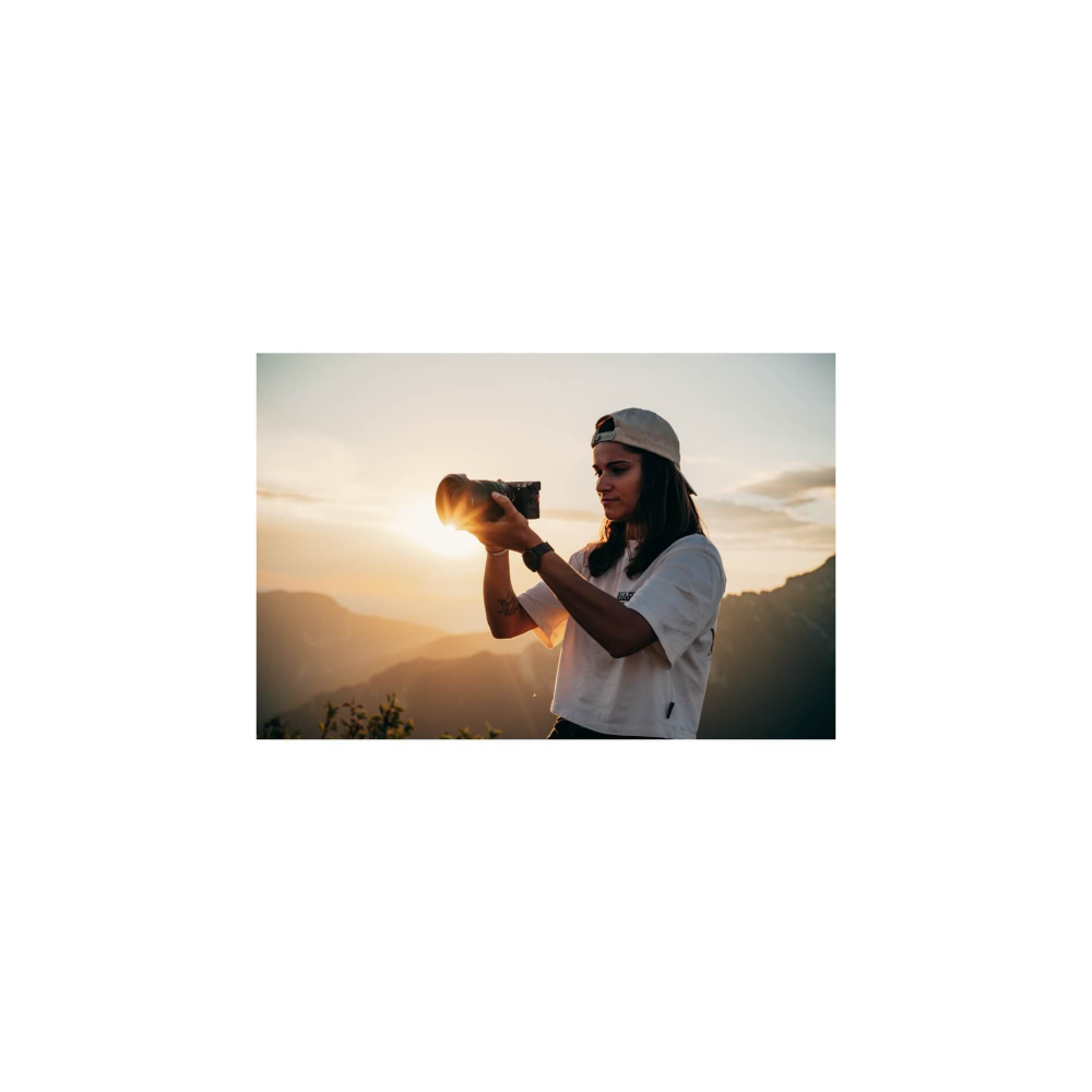 A woman outdoors holds a camera and takes a photo at sunset.