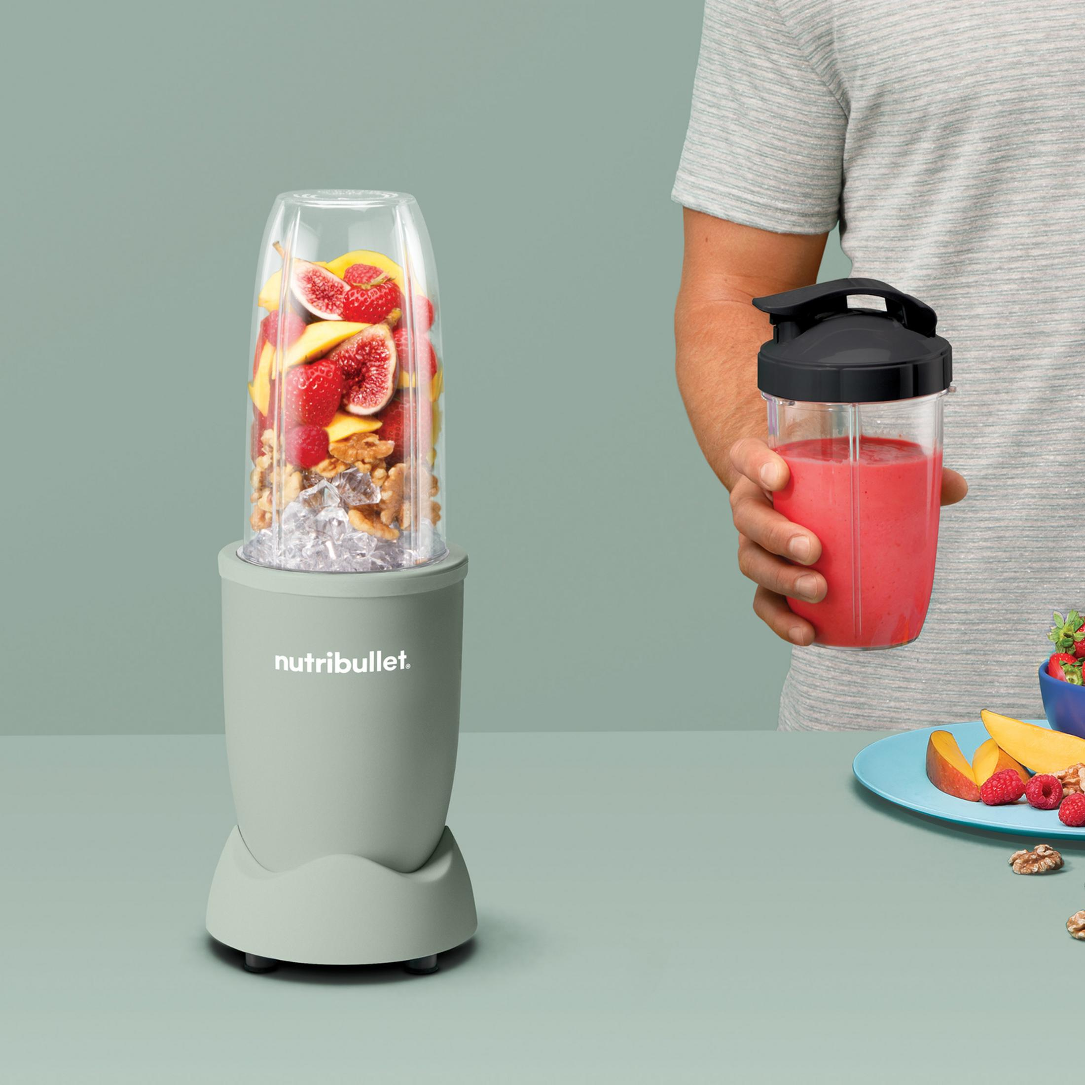A green blender with fruits and ice. Next to it, a man holds a finished smoothie.