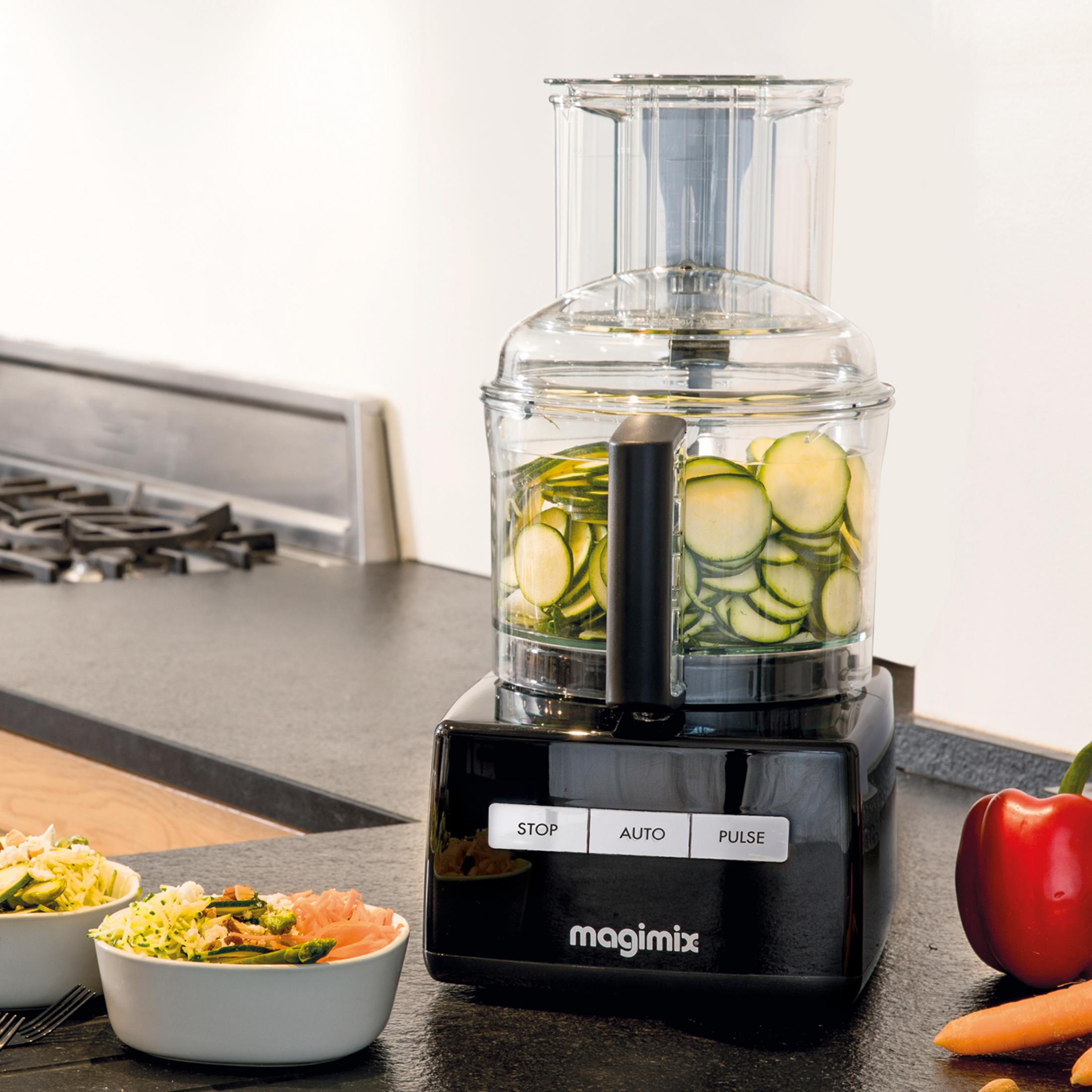 A black food processor with sliced zucchini in the container and bowls of salad.
