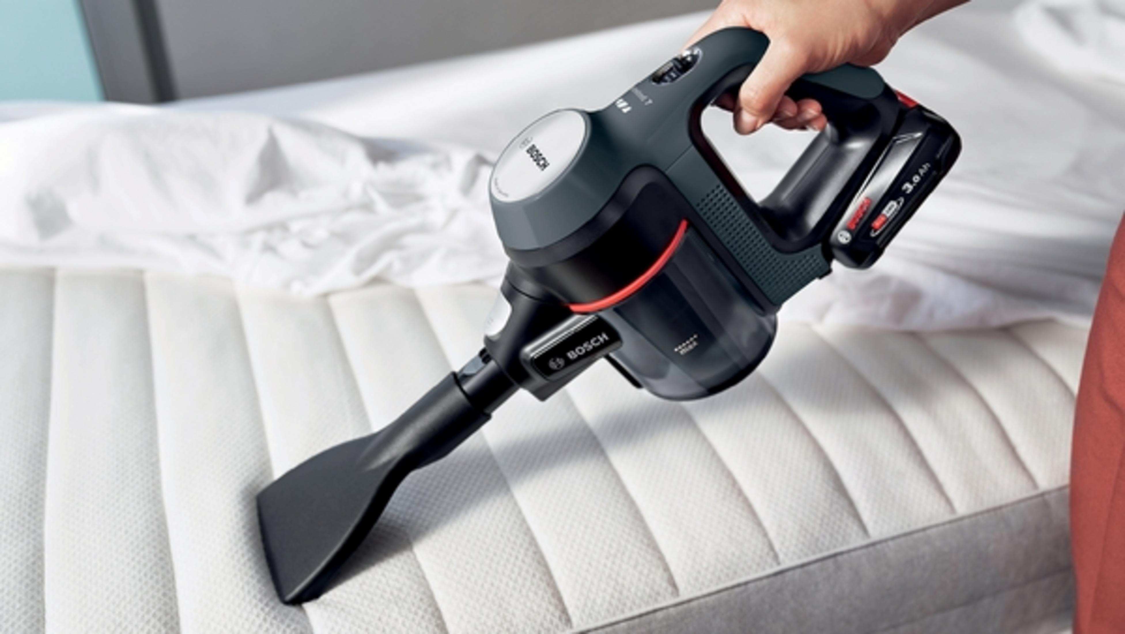Una mano tiene un aspirapolvere Bosch grigio scuro su un materasso bianco.