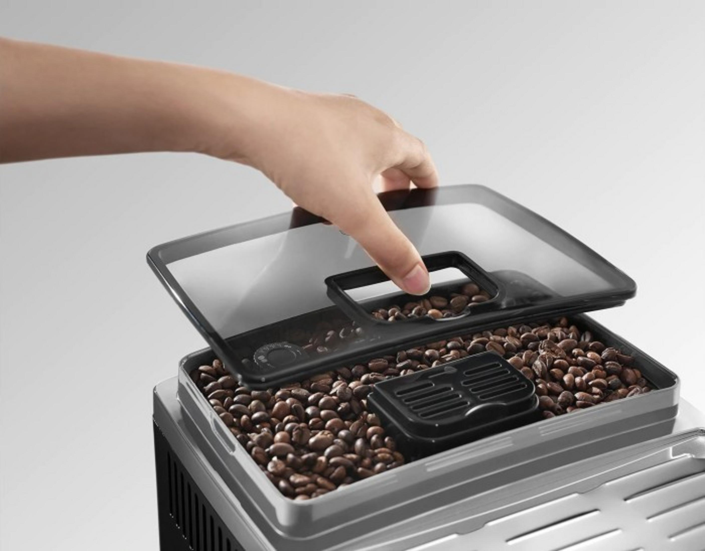 Una mano levanta una tapa transparente que revela granos de café en un molinillo.