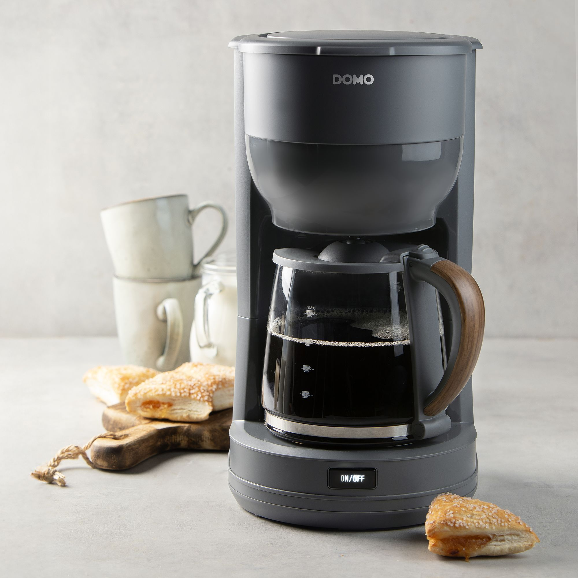 Grauer DOMO Kaffeemaschine mit Holzbalkenhenkel, gefüllter Kanne und Gebäck.