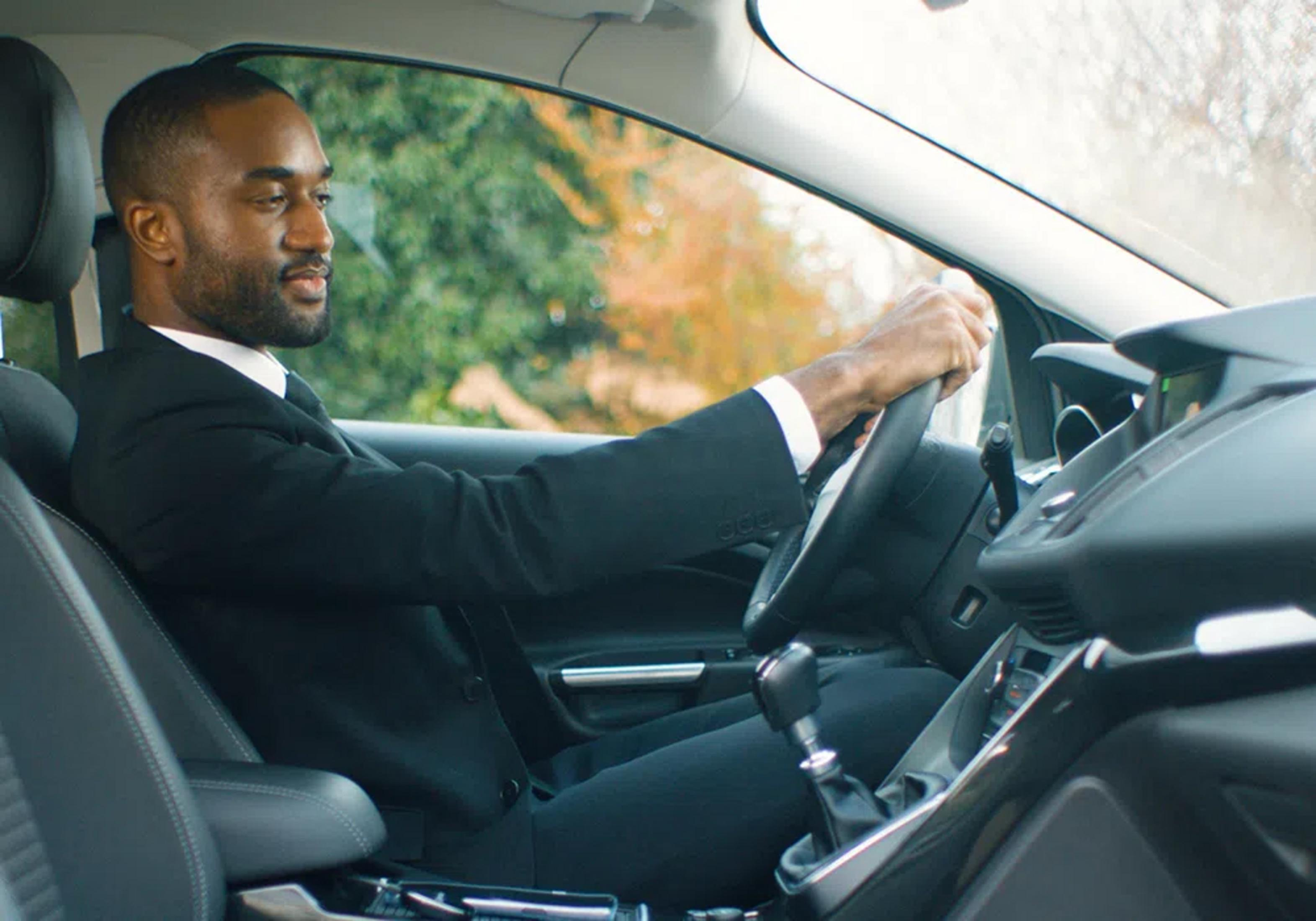 Un hombre con traje negro conduce un coche, manos en el volante.