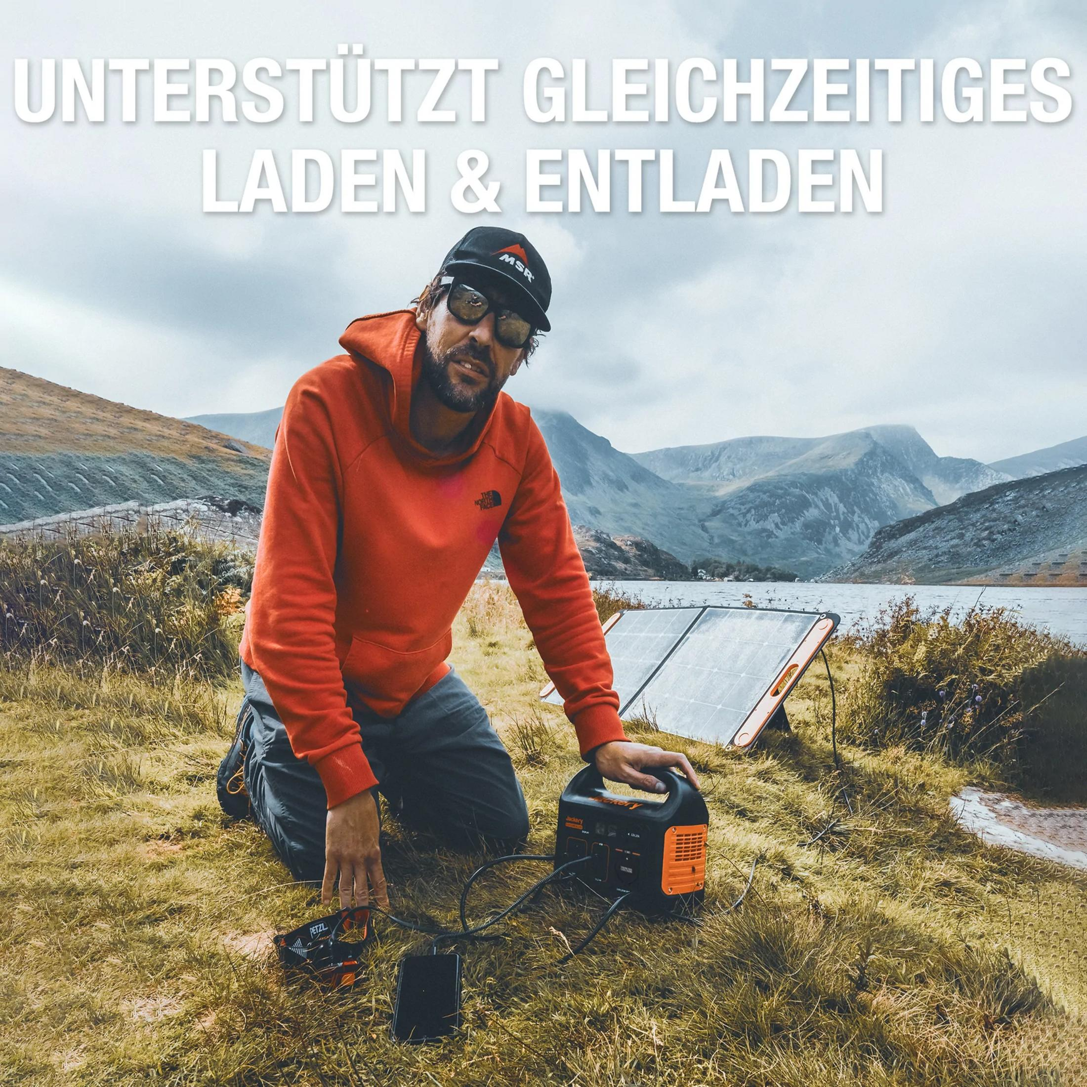 Man knielt in gras naast zonnepaneel, powerstation en telefoon. Bergen op achtergrond.