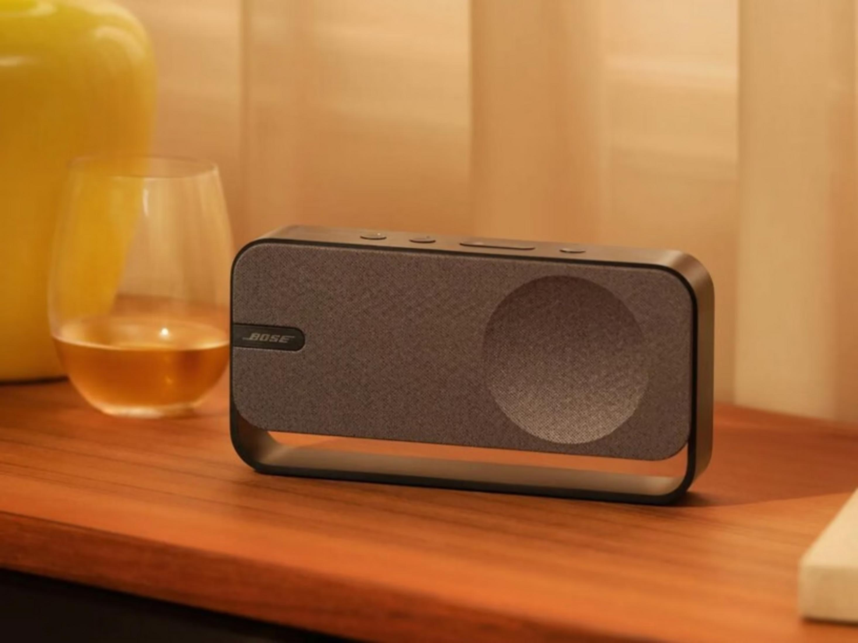 Un altavoz Bose gris se sienta sobre una mesa de madera junto a un vaso de líquido ámbar.