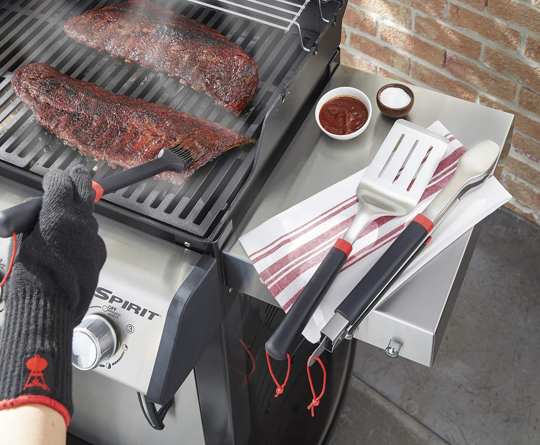 Costillas a la parrilla con salsa, sal, espátula, pinzas y cepillo en una parrilla Weber.