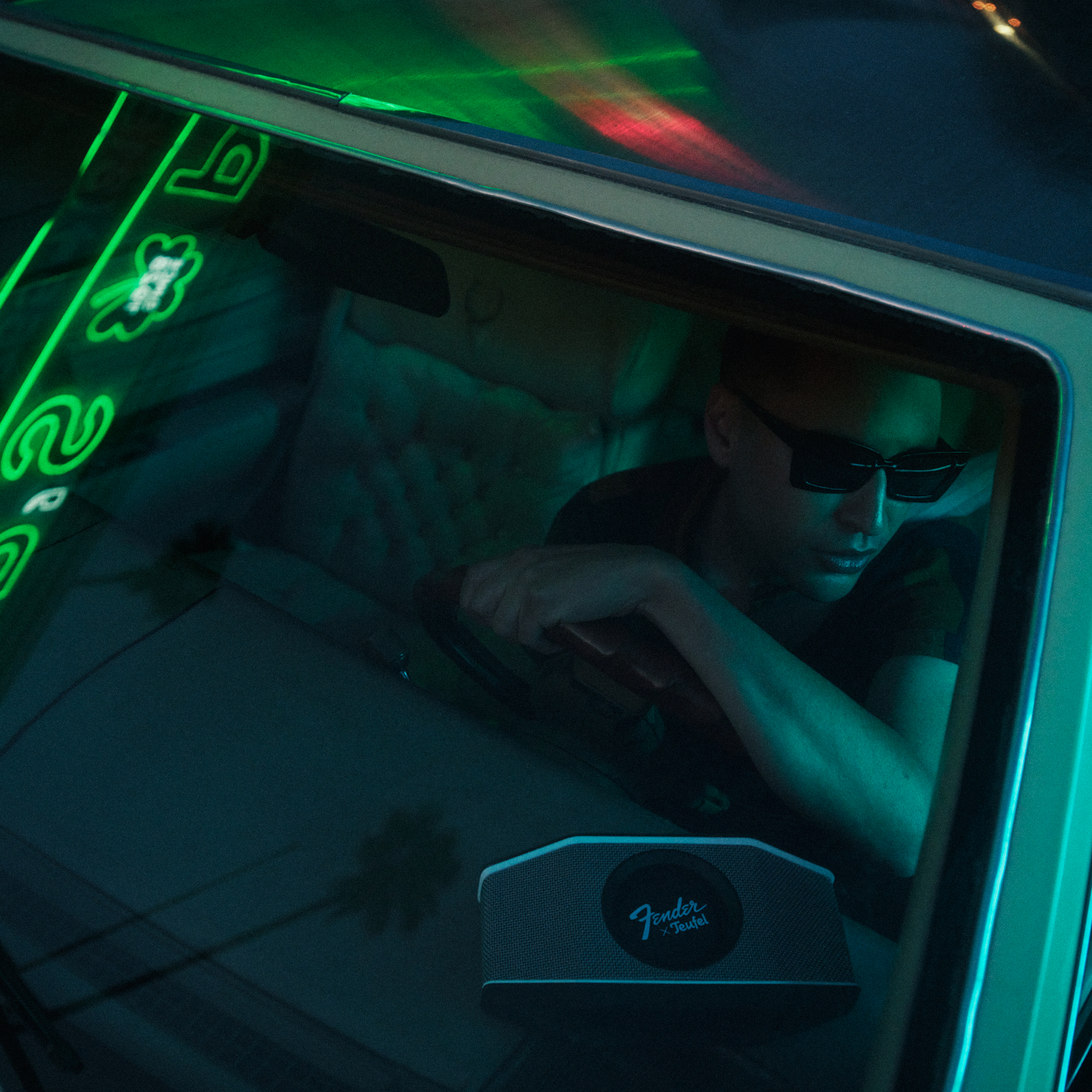 Mann mit Sonnenbrille fährt Auto, beleuchtet von grüner Neonwerbung.