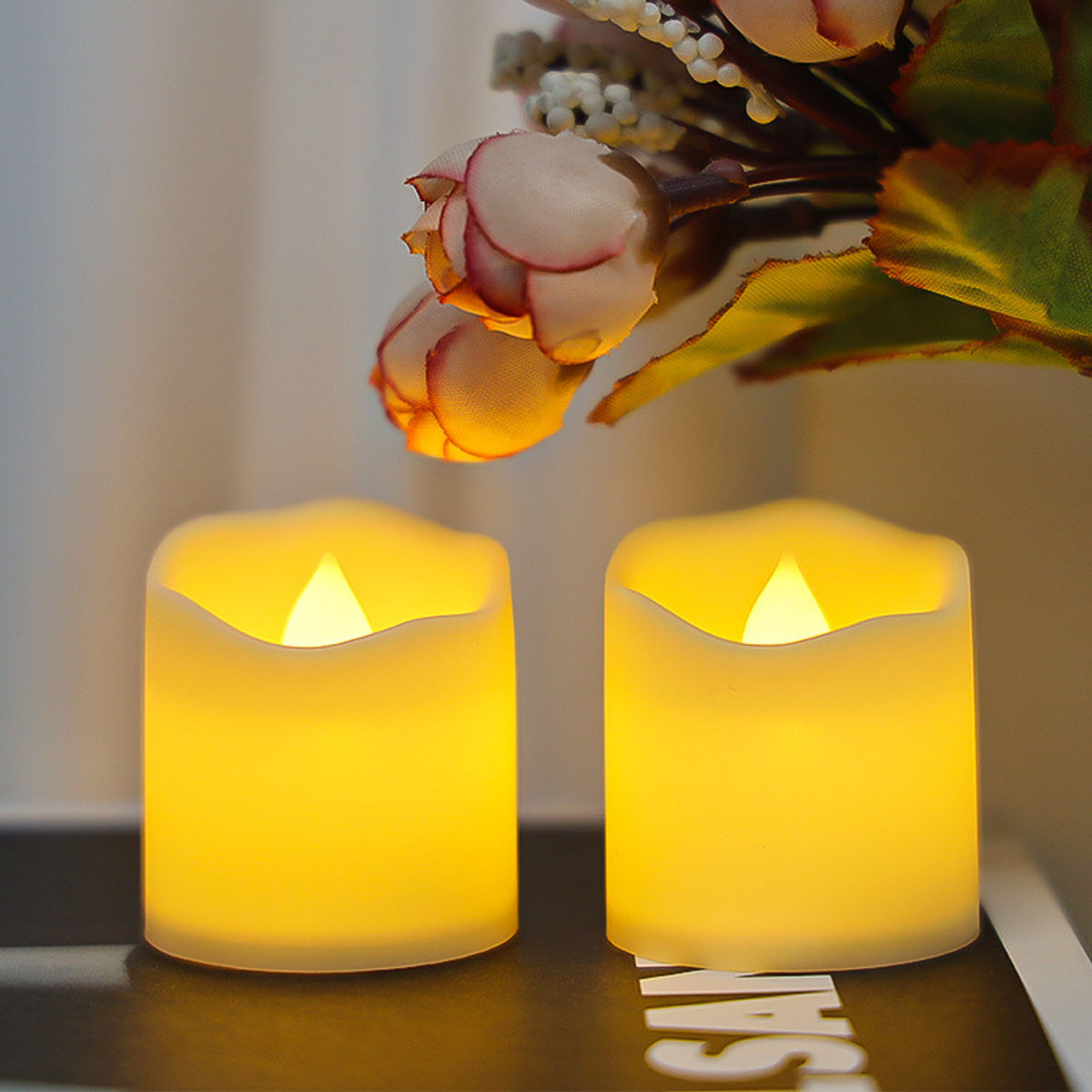 Zwei LED-Kerzen leuchten gelb auf einem dunklen Untergrund, im Hintergrund Blumen.