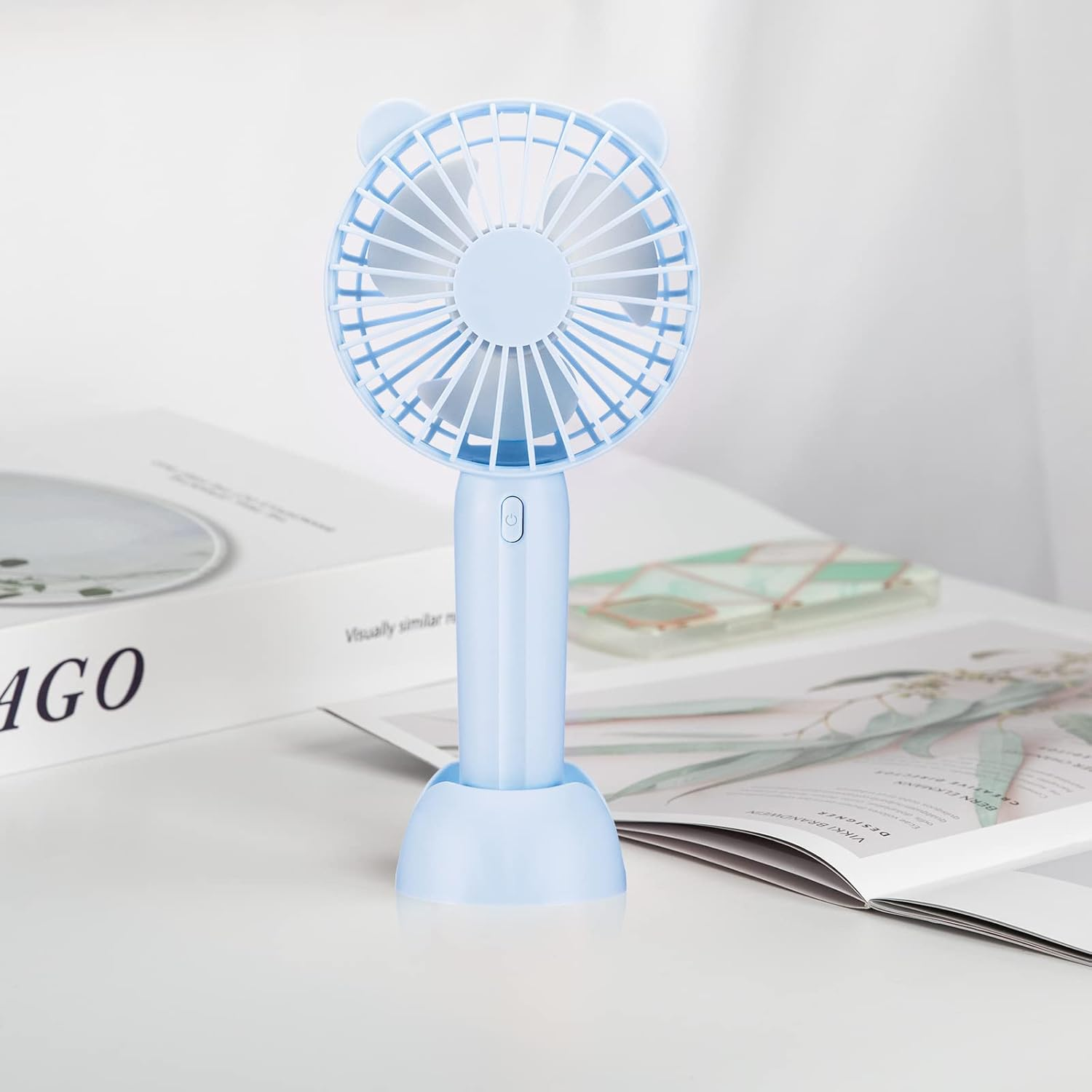 Ein hellblauer tragbarer Ventilator mit Bärenohren steht auf weißer Fläche neben offenen Büchern.