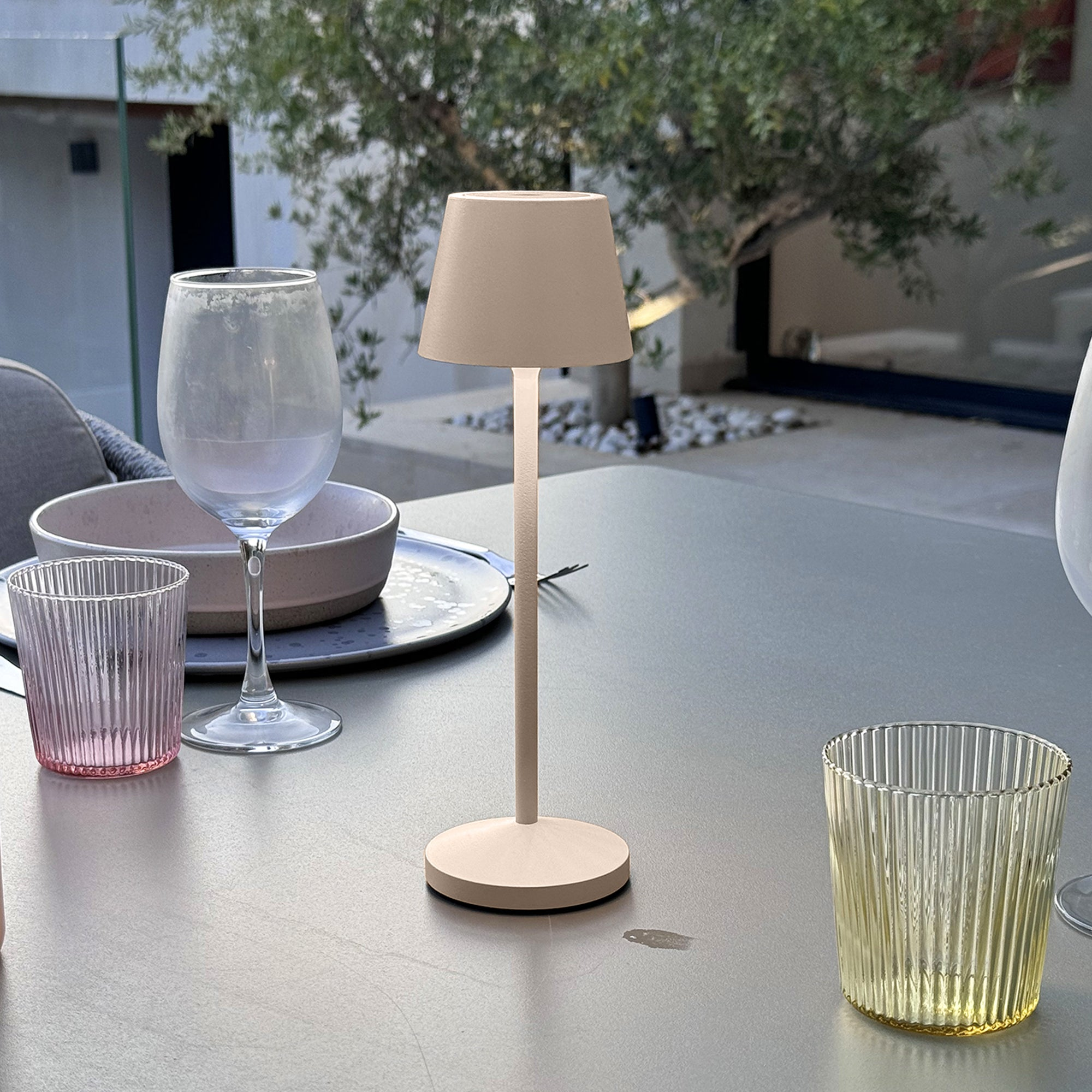 Ein heller Tisch mit Gläsern und einer Lampe, im Hintergrund ein Baum.