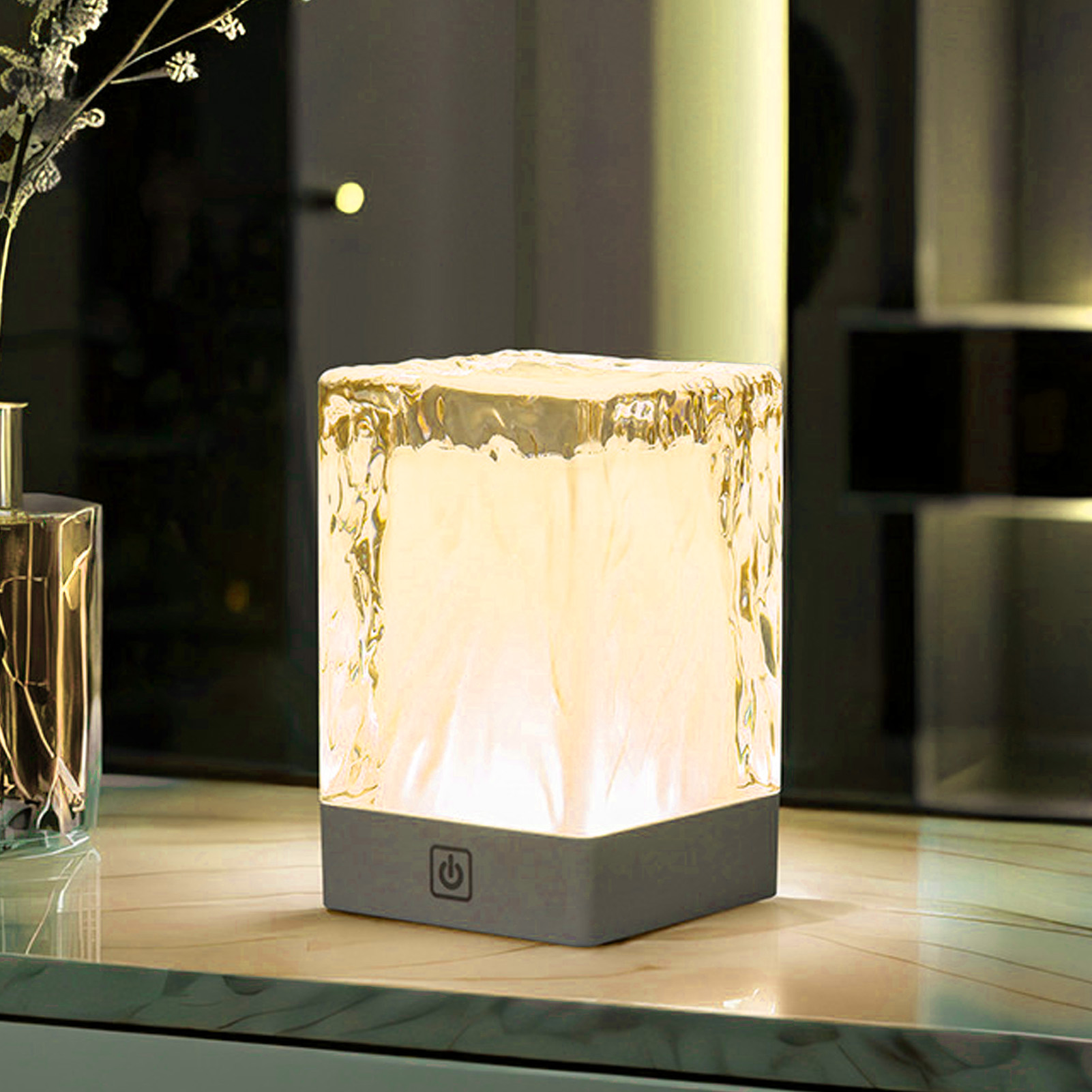 Ein würfelförmiges Lichtobjekt mit Eisstruktur leuchtet gelblich. Es steht auf einem Tisch neben einer Vase.