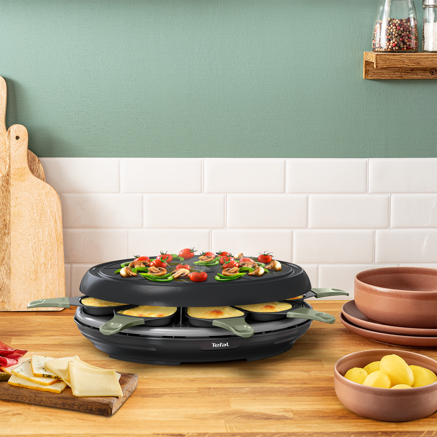 Una griglia per raclette con formaggio e verdure in cima, accanto a formaggio e patate.