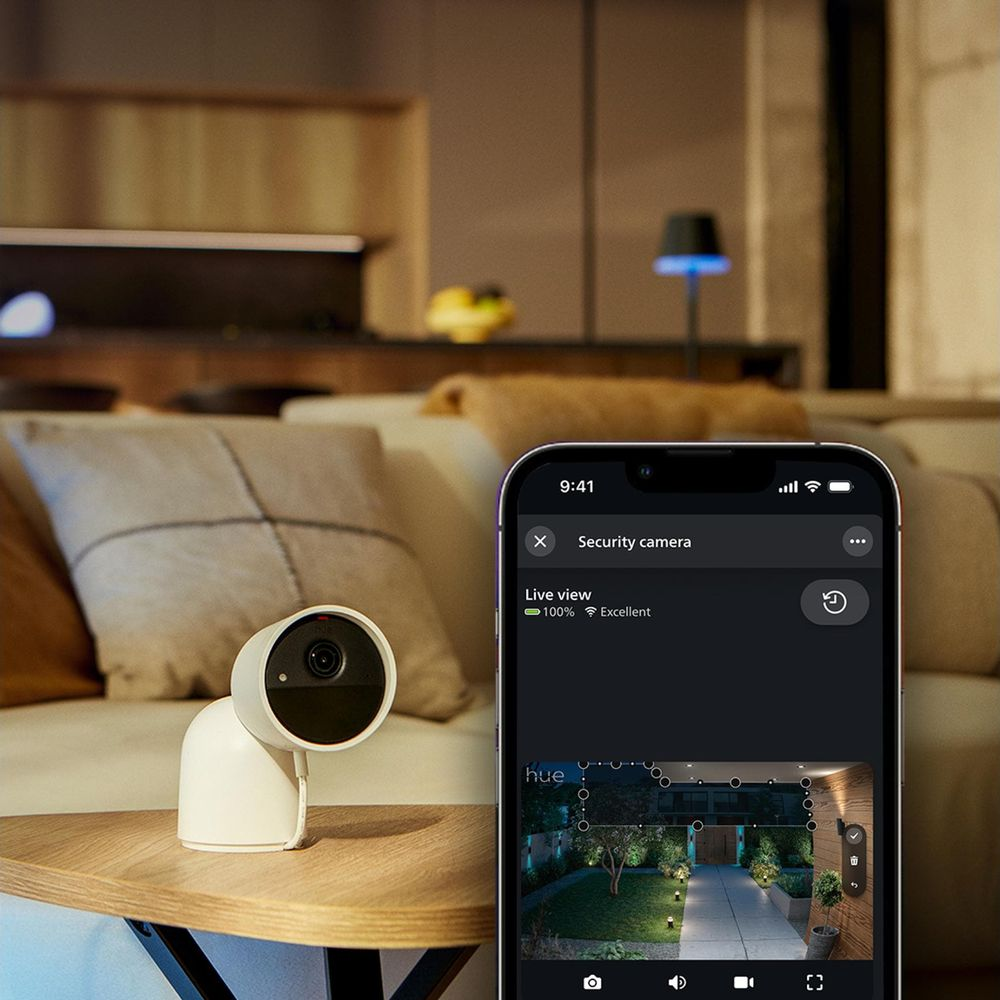 Eine weiße Sicherheitskamera steht auf einem Holztisch neben einem Handy mit Live-Ansicht.