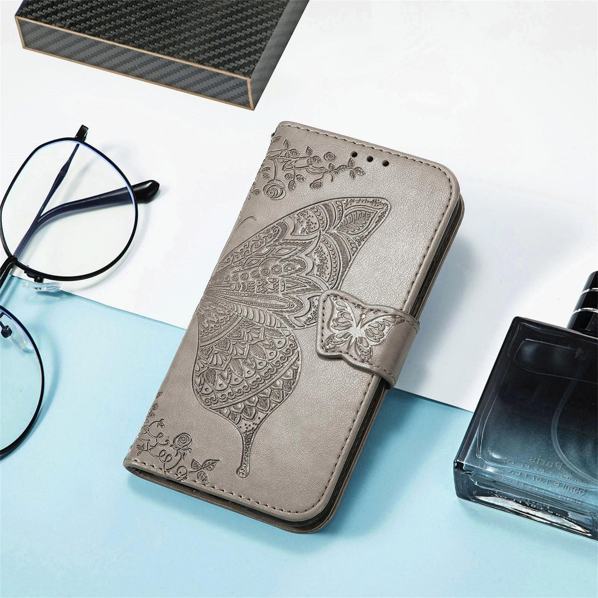 Graues Handycover mit Schmetterlingsprägung, Brille, Parfüm und Carbon-Box.