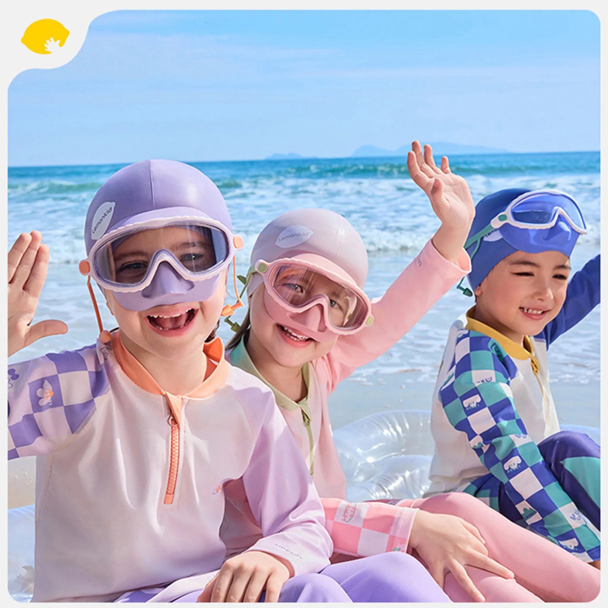 Drei Kinder in Badeanzügen und Schwimmbrillen sitzen am Strand. Das Meer und der Himmel sind im Hintergrund.