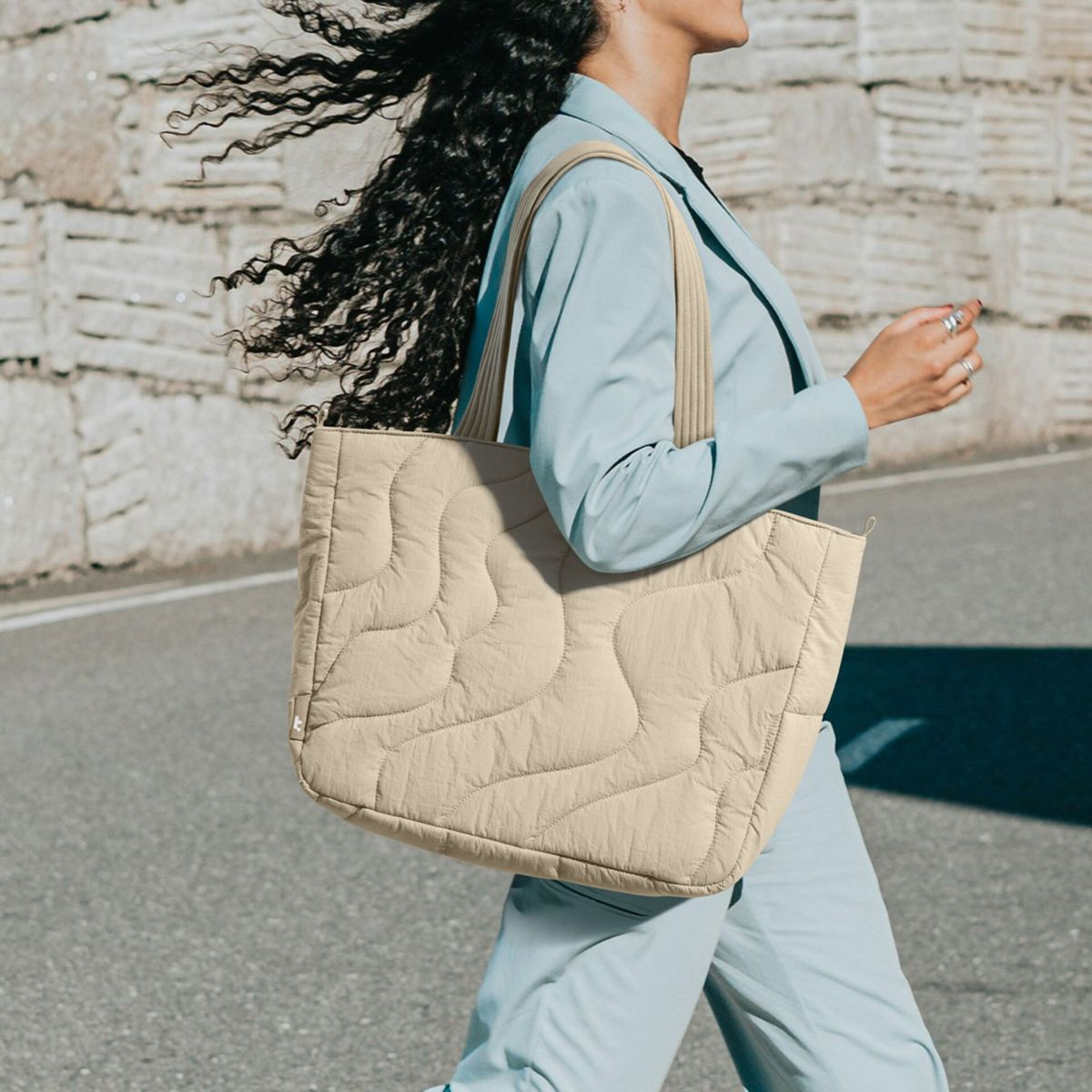 Una donna con lunghi capelli neri indossa una giacca azzurro chiaro e una borsa shopper trapuntata beige.