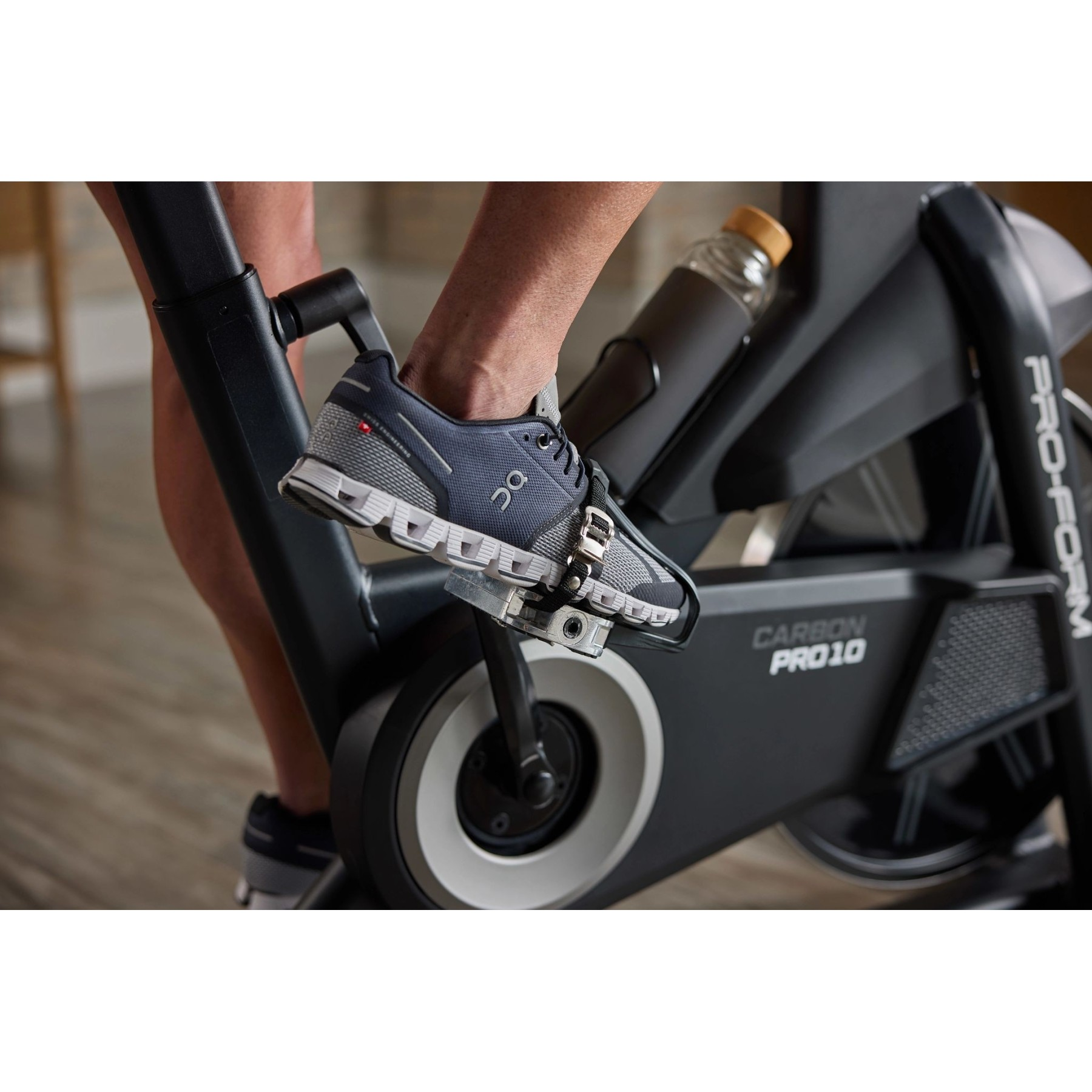 Ein Mann tritt auf einem Indoor-Bike in die Pedale, trägt dunkle Turnschuhe und hat eine Wasserflasche dabei.