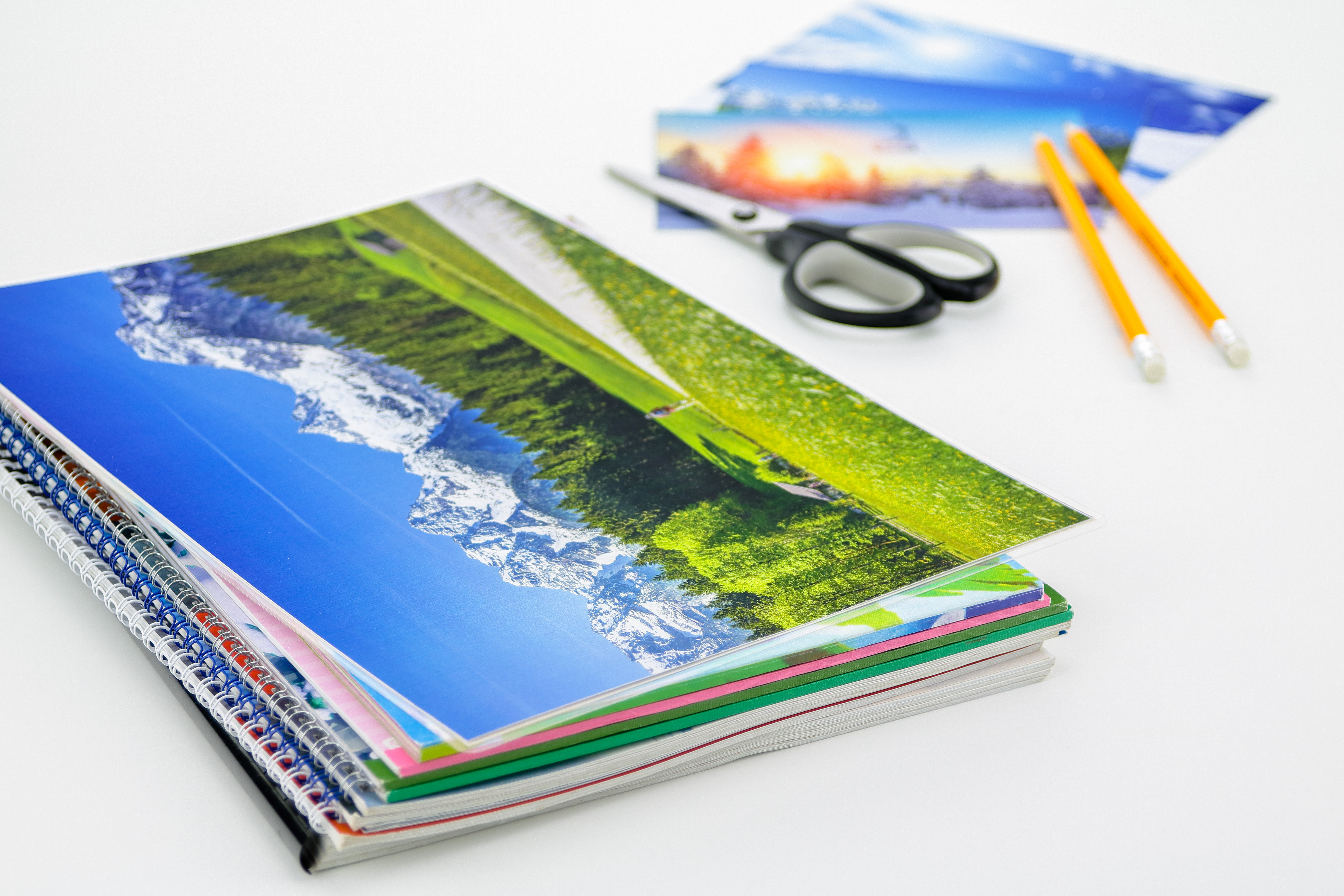 Pile de carnets avec photos de paysages, ciseaux et crayons sur fond blanc.