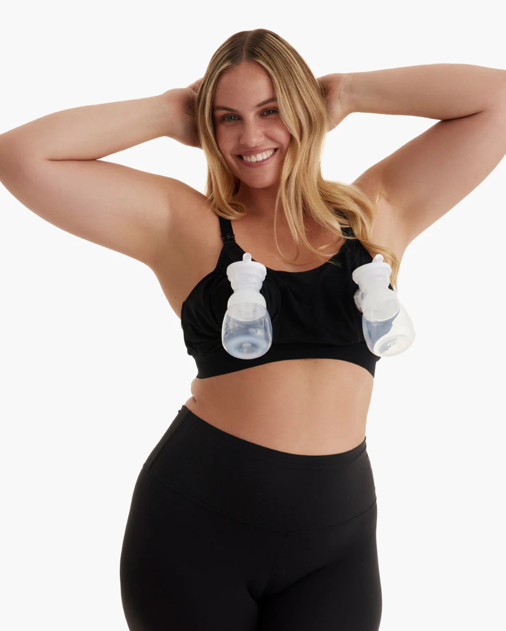 Mujer con cabello rubio lleva sujetador y leggings negros, manos tras la cabeza.