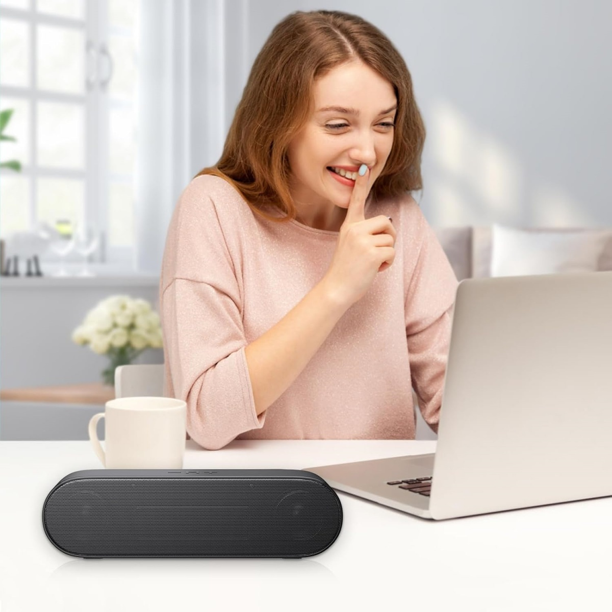 Eine lächelnde Frau mit Finger auf den Lippen sitzt an einem Tisch mit Laptop, Lautsprecher und Becher.