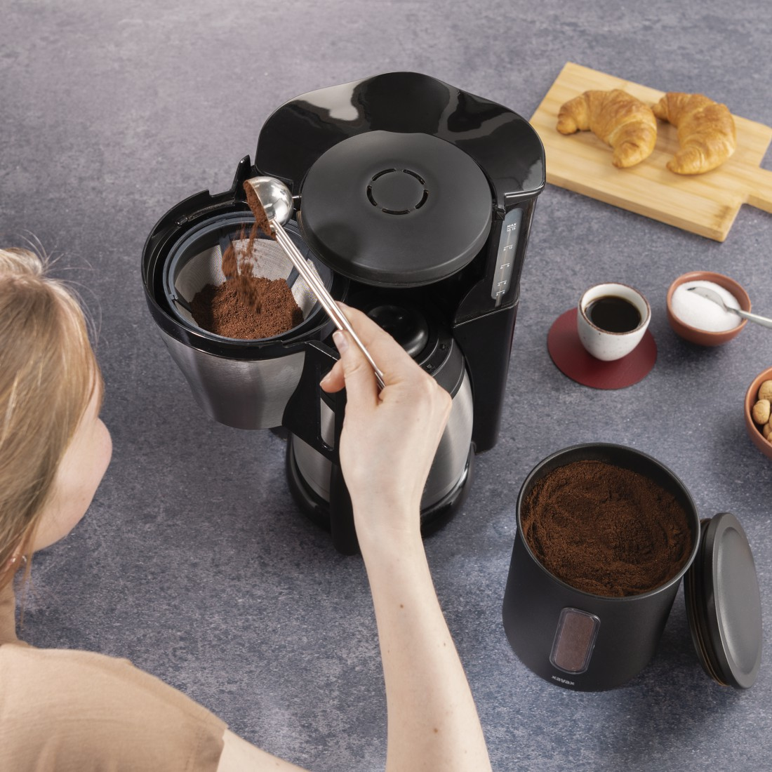 Eine Frau gibt Kaffeepulver in eine Kaffeemaschine. Daneben stehen Croissants, Kaffee, Zucker und Nüsse.
