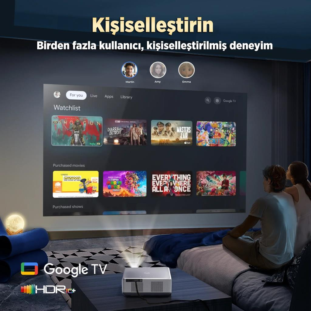 Projektör, film ve profillerle Google TV arayüzünü gösteriyor. İki kişi yatakta oturuyor.