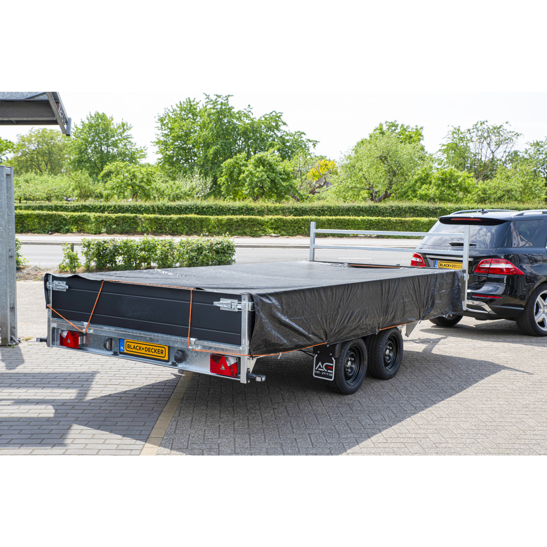 Zwarte trailer met zeil gekoppeld aan een zwarte SUV op een verhard terrein.