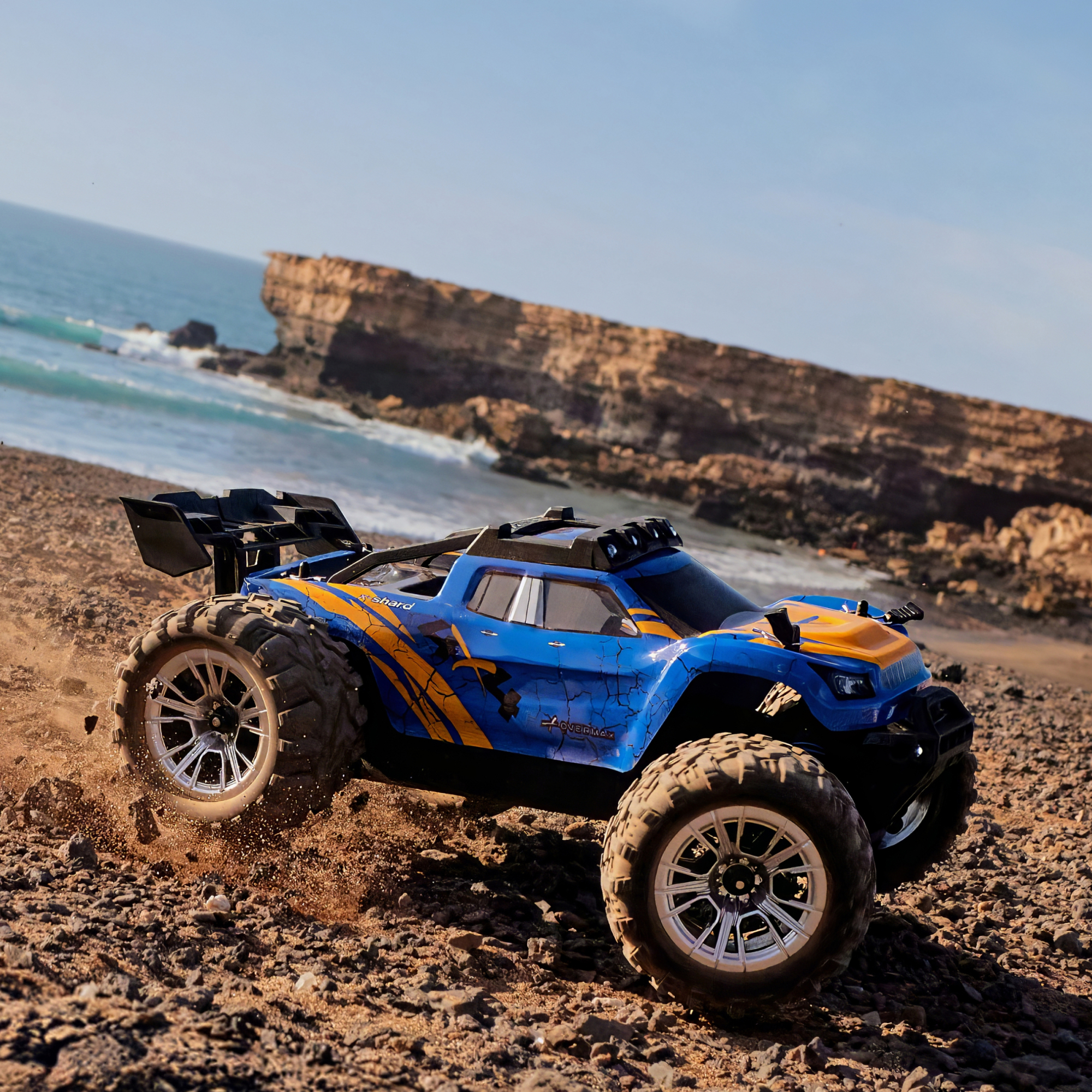 Blau-oranger RC-Buggy fährt auf felsigem Strand neben dem Meer.