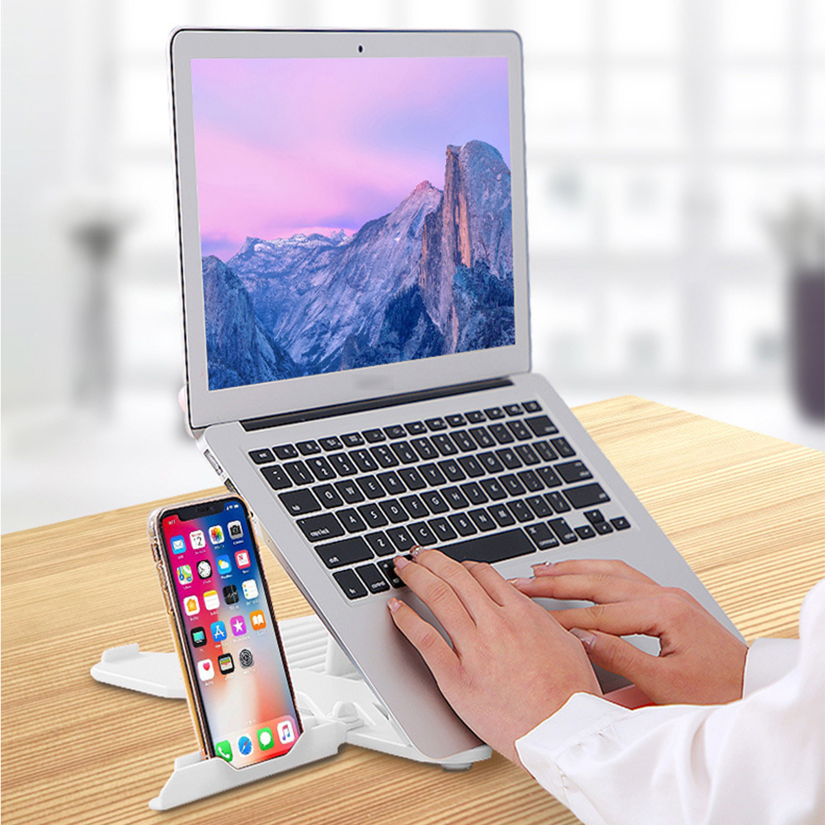 Hände tippen auf einem silbernen Laptop auf einem weißen Ständer, daneben ein Telefon auf einem Ständer.