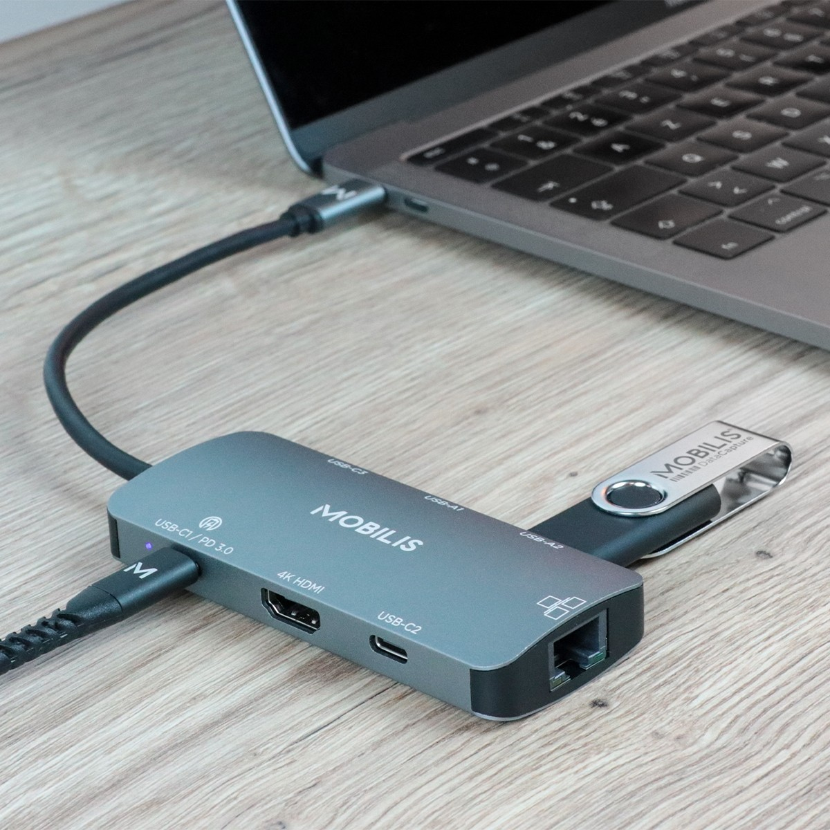 Ein grauer USB-C-Hub mit Kabel, einem USB-Stick und einem Laptop auf einem Holztisch.