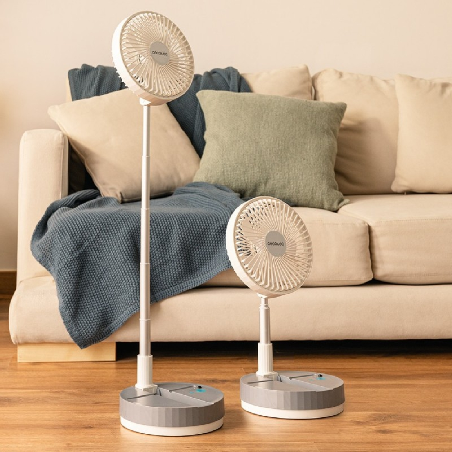 Dos ventiladores portátiles blancos sobre suelo de madera frente a un sofá.