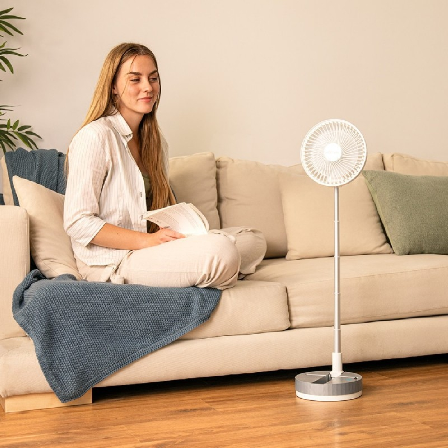 Mujer leyendo en sofá junto a ventilador blanco en suelo de madera.