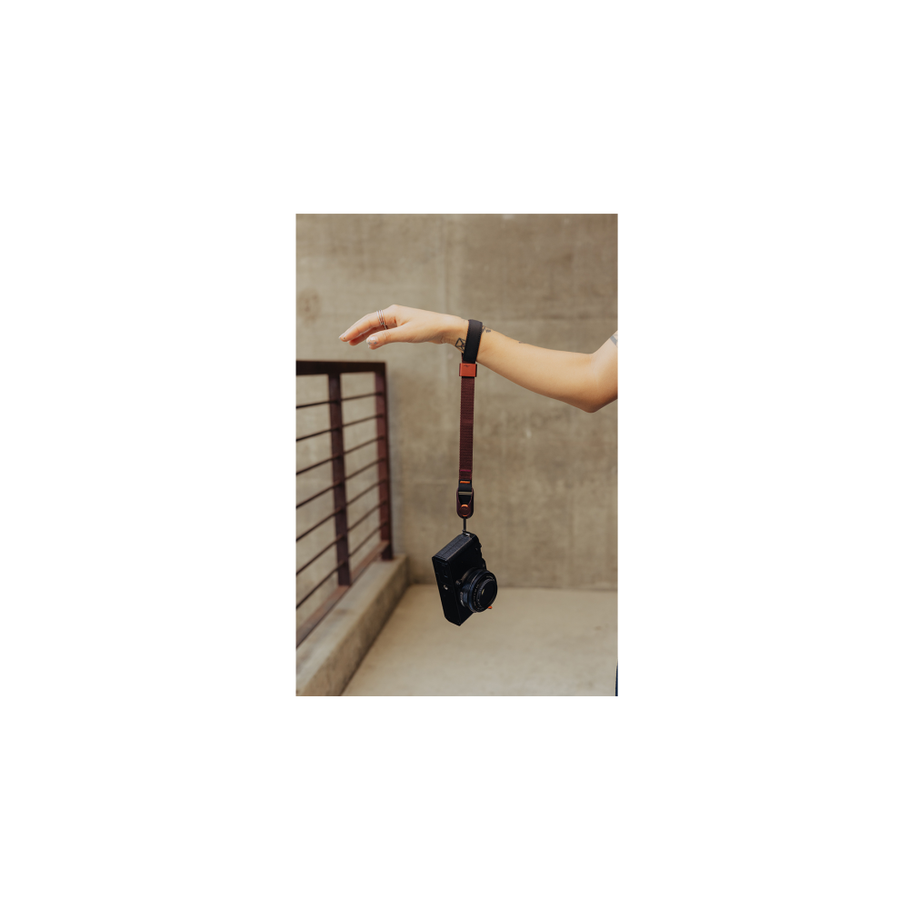 A hand holds a black camera on a brown wrist strap. Concrete wall in the background.