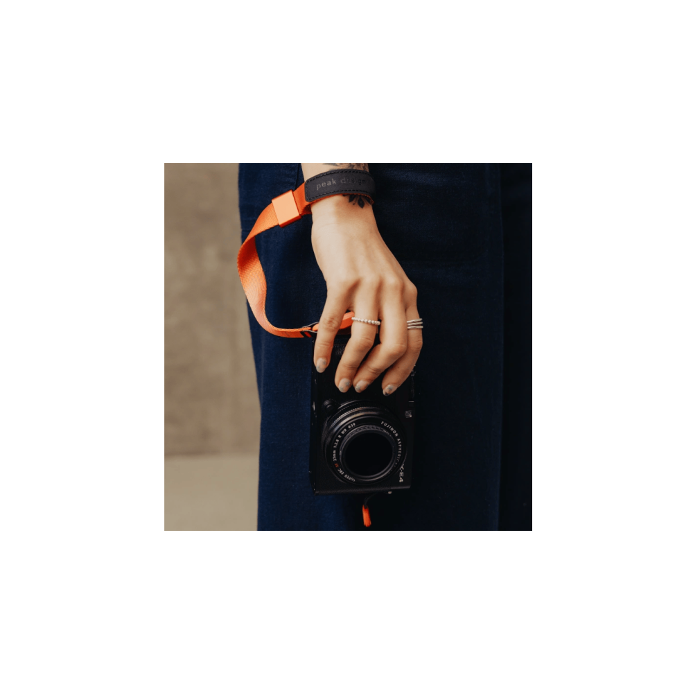 Een hand met een oranje band houdt een zwarte camera vast. Er worden ringen gedragen.