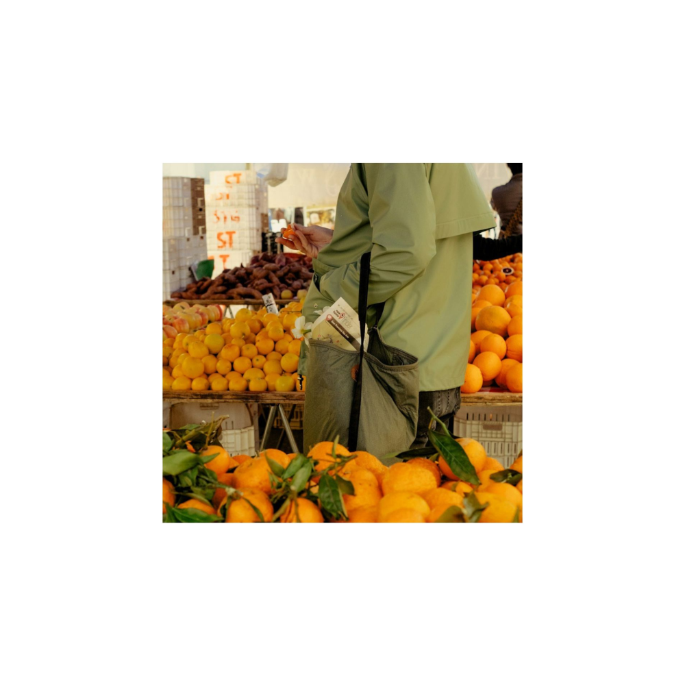 Een persoon in een groen jasje houdt een sinaasappel vast bij een marktkraam vol fruit.