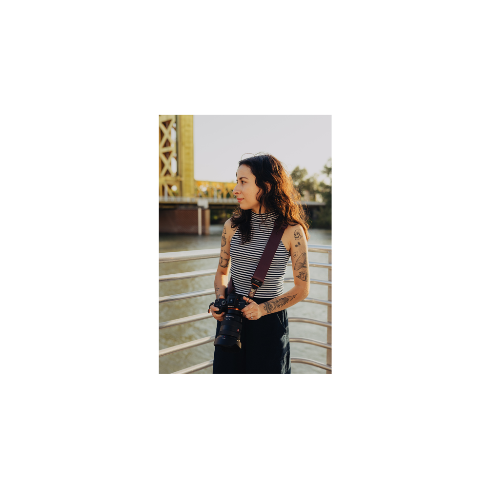 Een vrouw met tatoeages houdt een camera vast, staand voor een brug.
