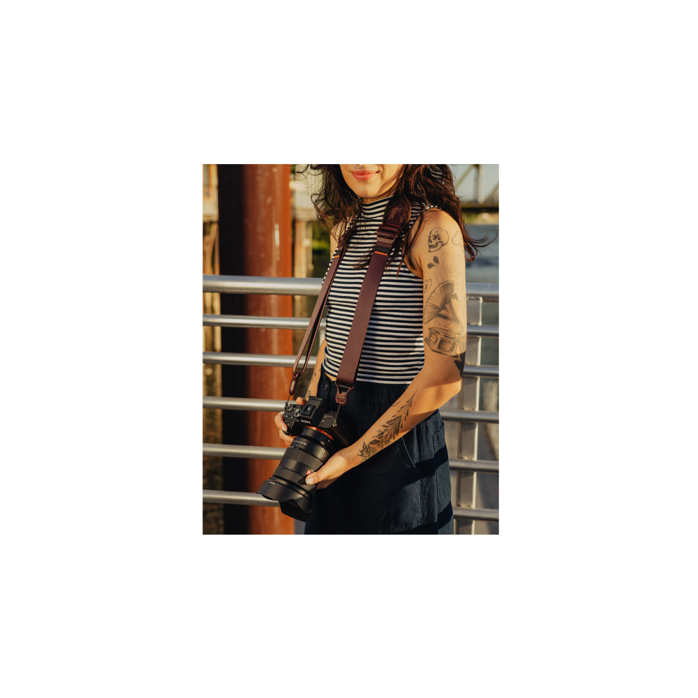 Getatoeëerde vrouw met camera en riem, gestreept topje en donkere broek.