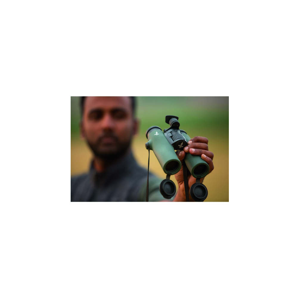 A man holds green binoculars with a Swarovski logo. Blurred background.