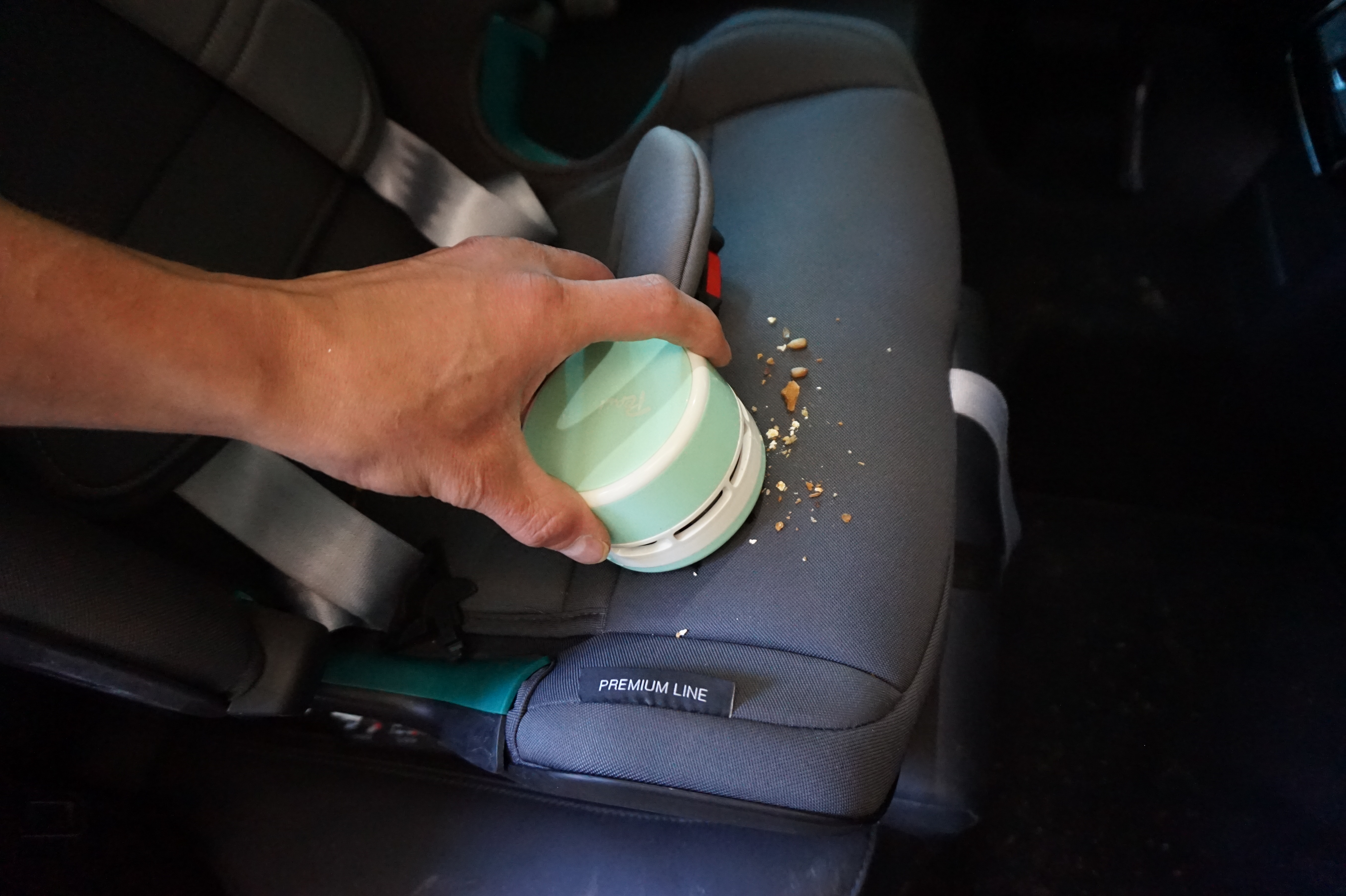 Hand hält einen kleinen mintgrünen Staubsauger auf einem Autositz mit Krümeln.