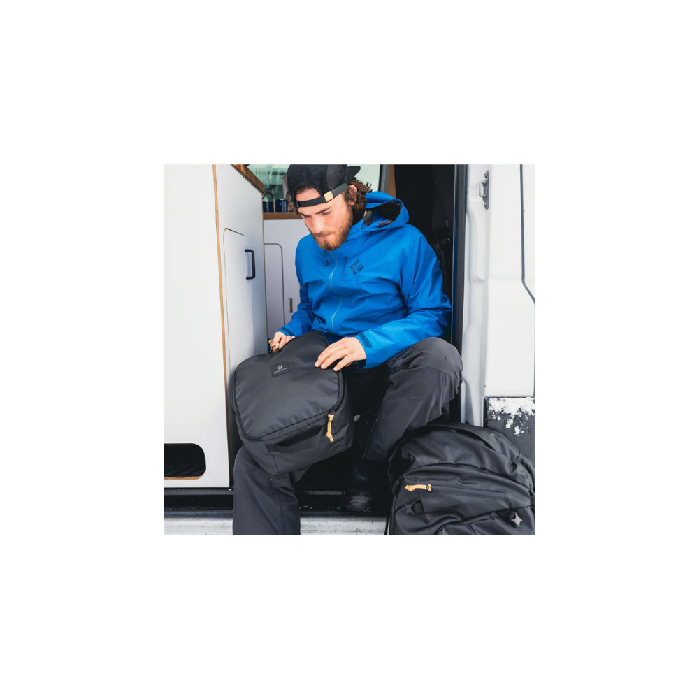 Man in blue jacket sits in van, packing black backpack. Another backpack is beside him.