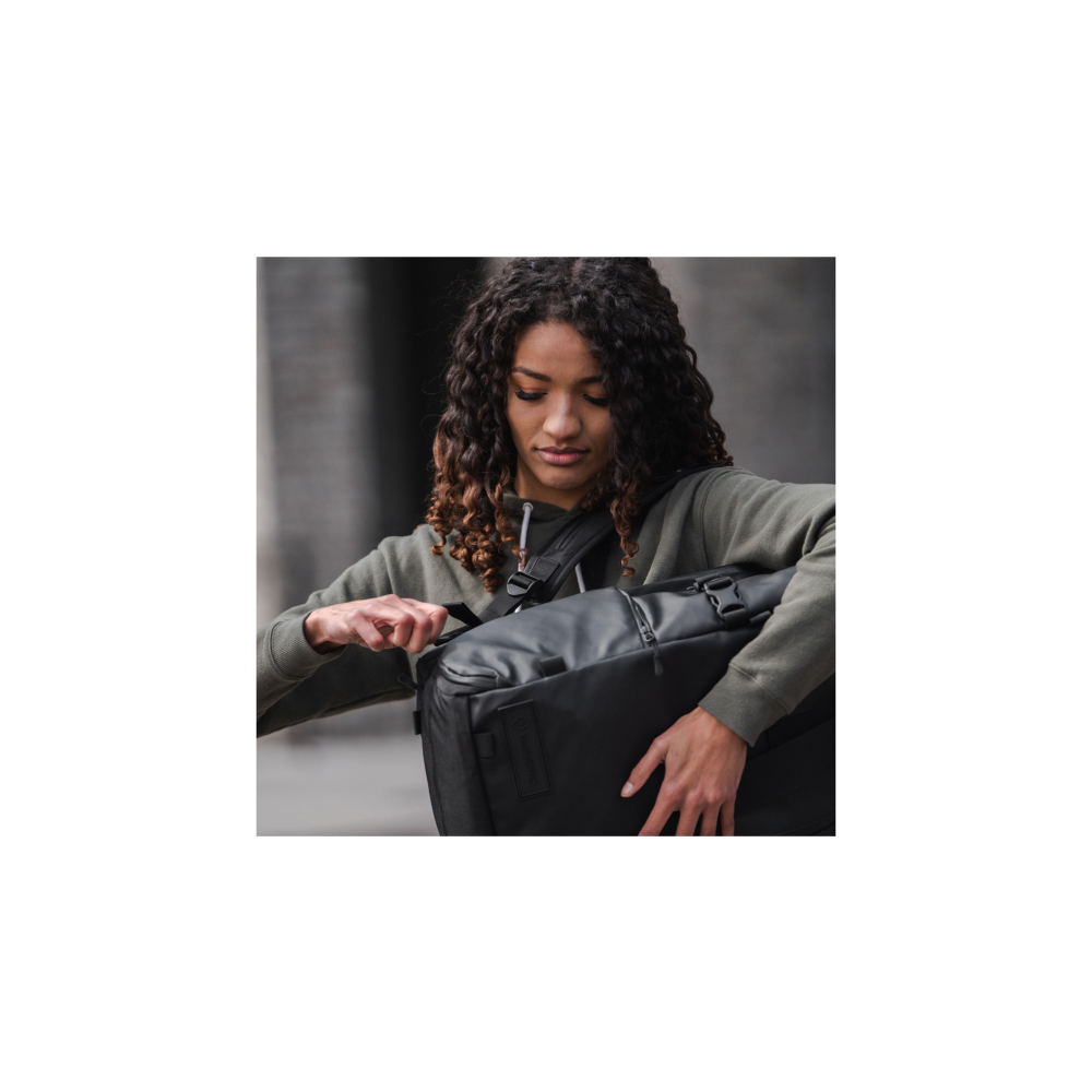Een vrouw met krullend haar in een groene hoodie opent een zwarte tas.