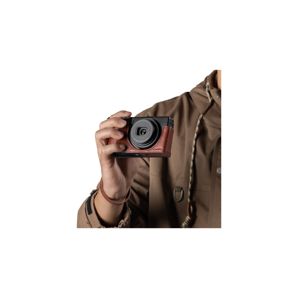A hand holds a black camera with a brown leather case. The person wears a brown jacket.